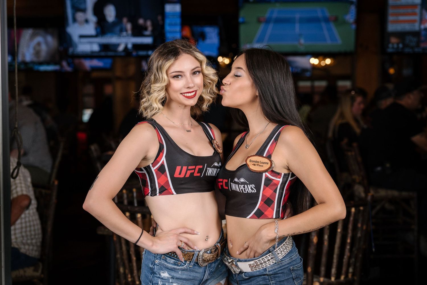 Two models in UFC tops and denim shorts pose together at sports bar.