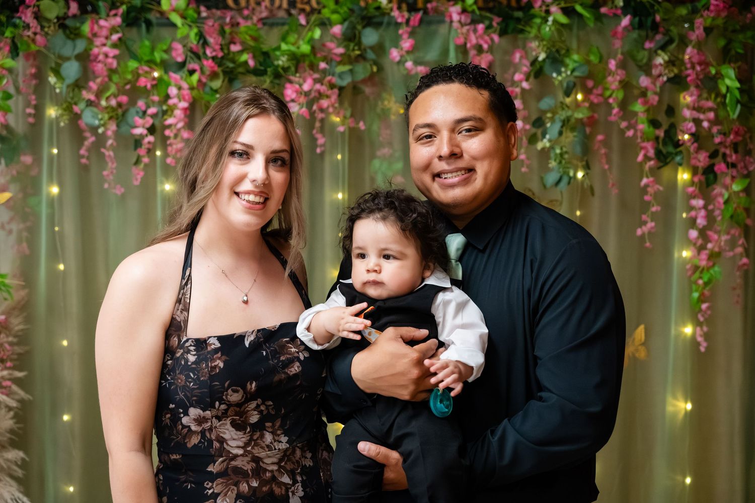 A smiling couple poses with their baby boy in front of a floral backdrop with greenery and fairy lights.