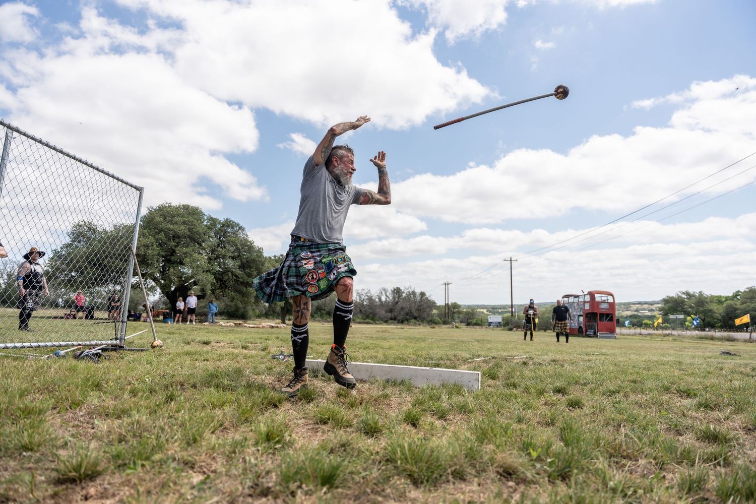 Man in kilt throwing hammer at Highland Games competition outdoors on green field under partly cloudy sky.