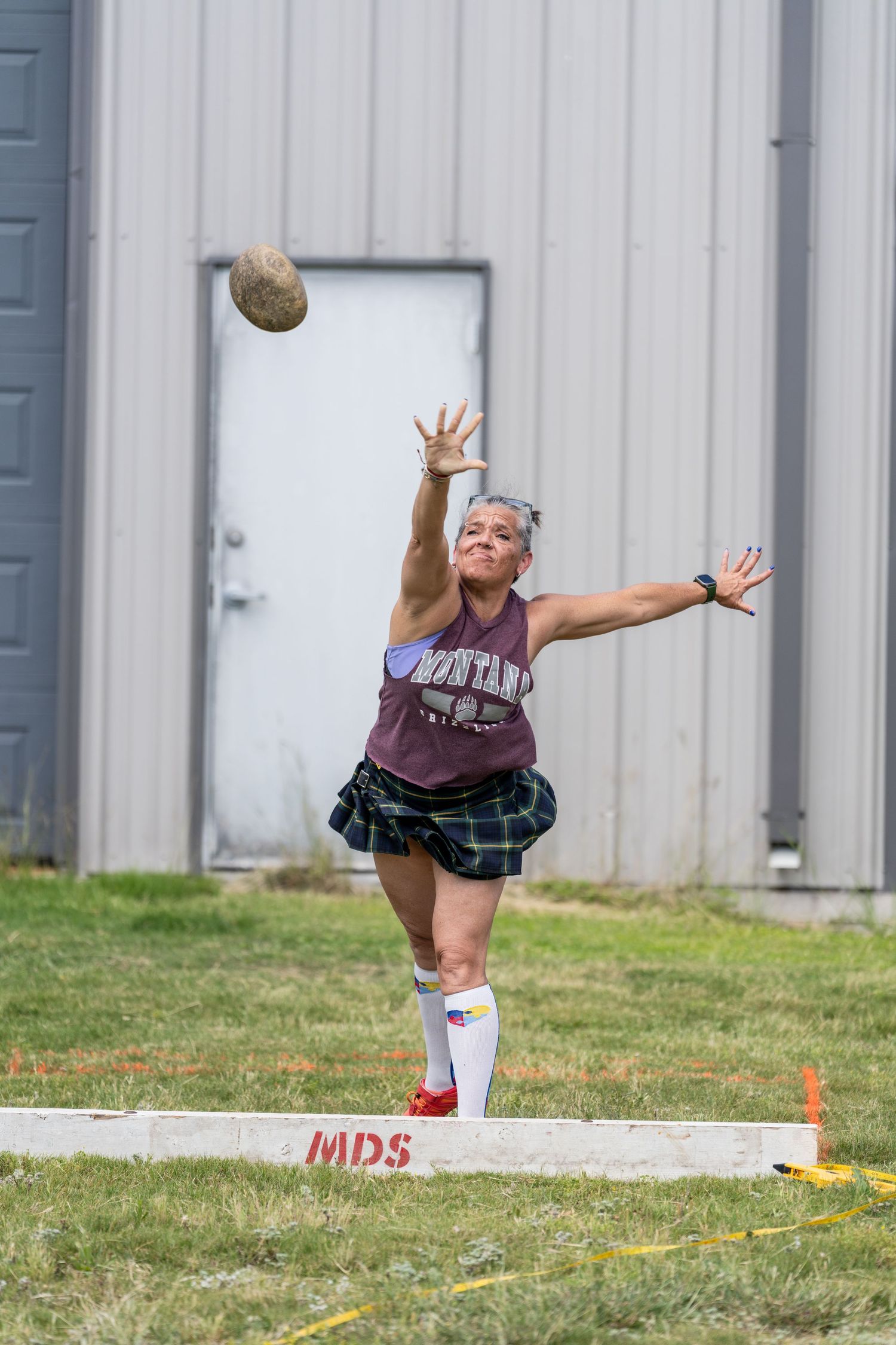 Woman in plaid skirt throwing a stone at a Highland Games competition outdoors near a metal building.