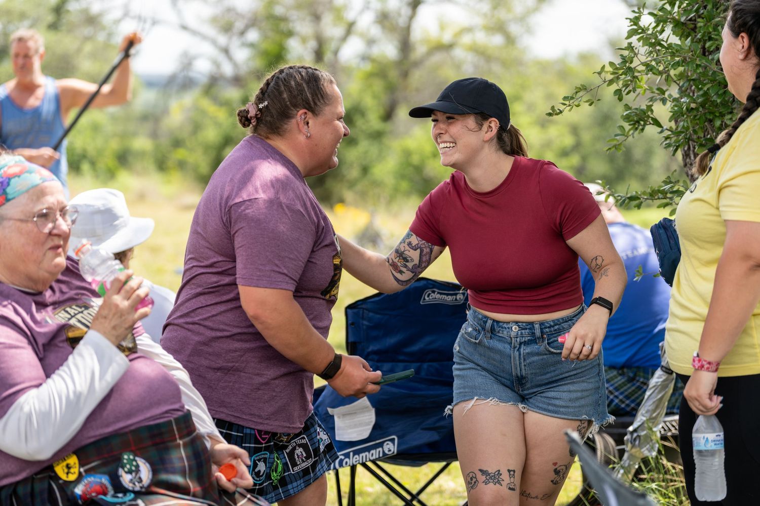 Two women greeting each other warmly at an outdoor gathering with others seated nearby on a sunny day.