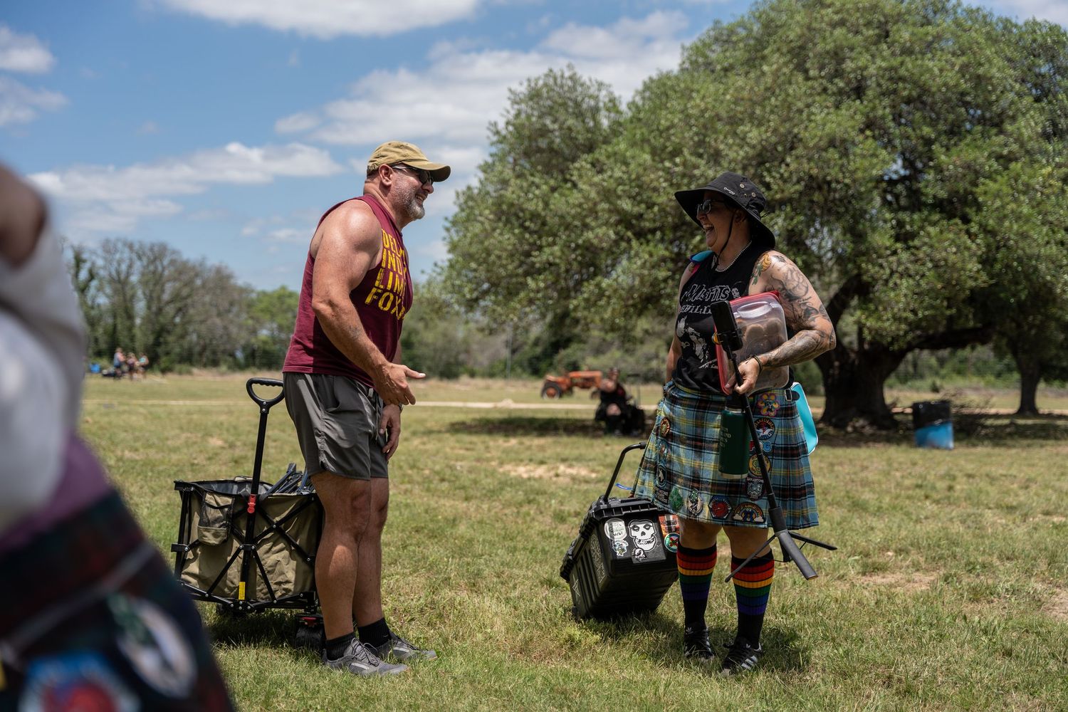 Two people in casual outdoor attire chat in a sunny green field, carrying bags near large shade trees.