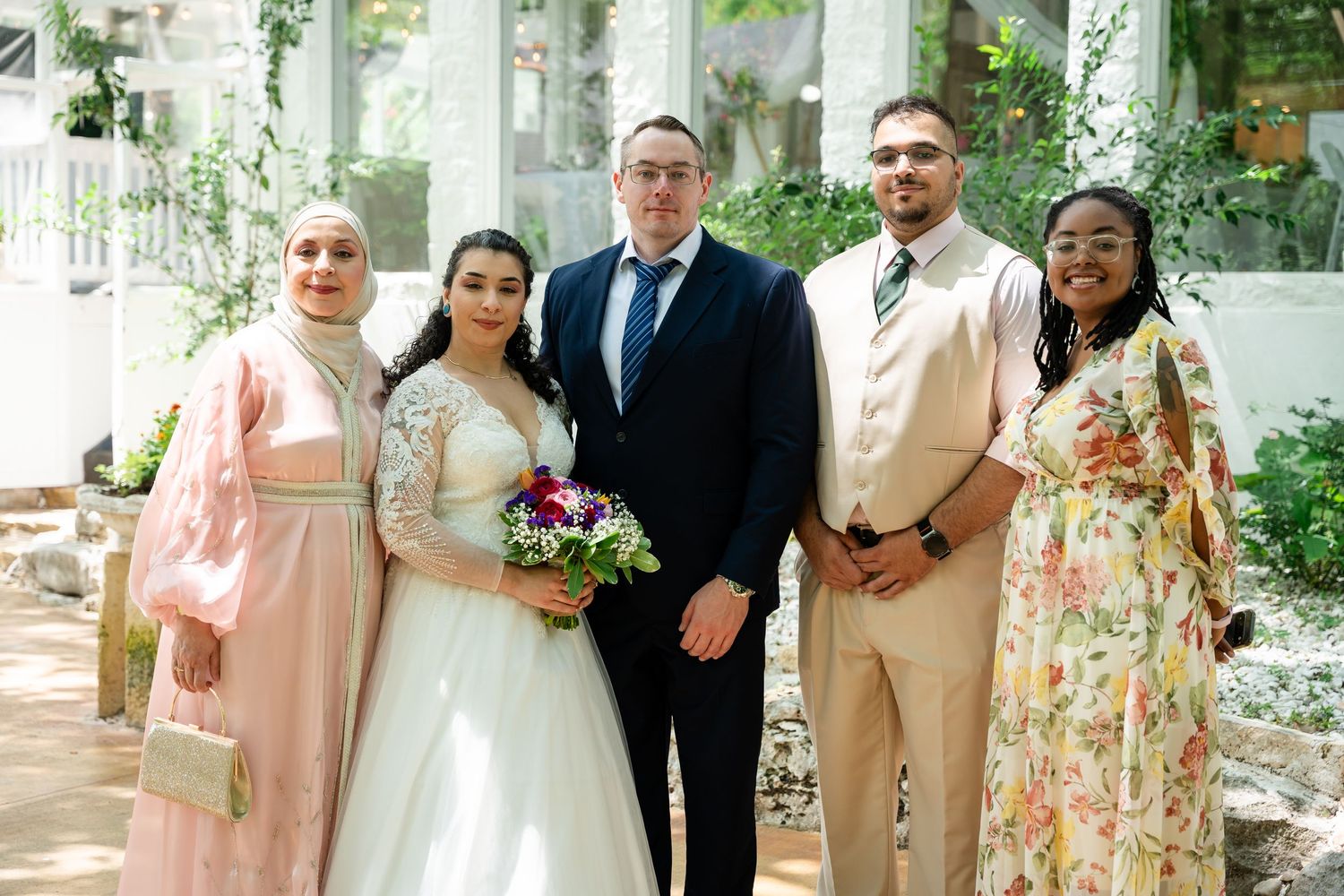 Wedding party of five posing indoors, bride in white gown holding bouquet, guests in formal and floral attire.