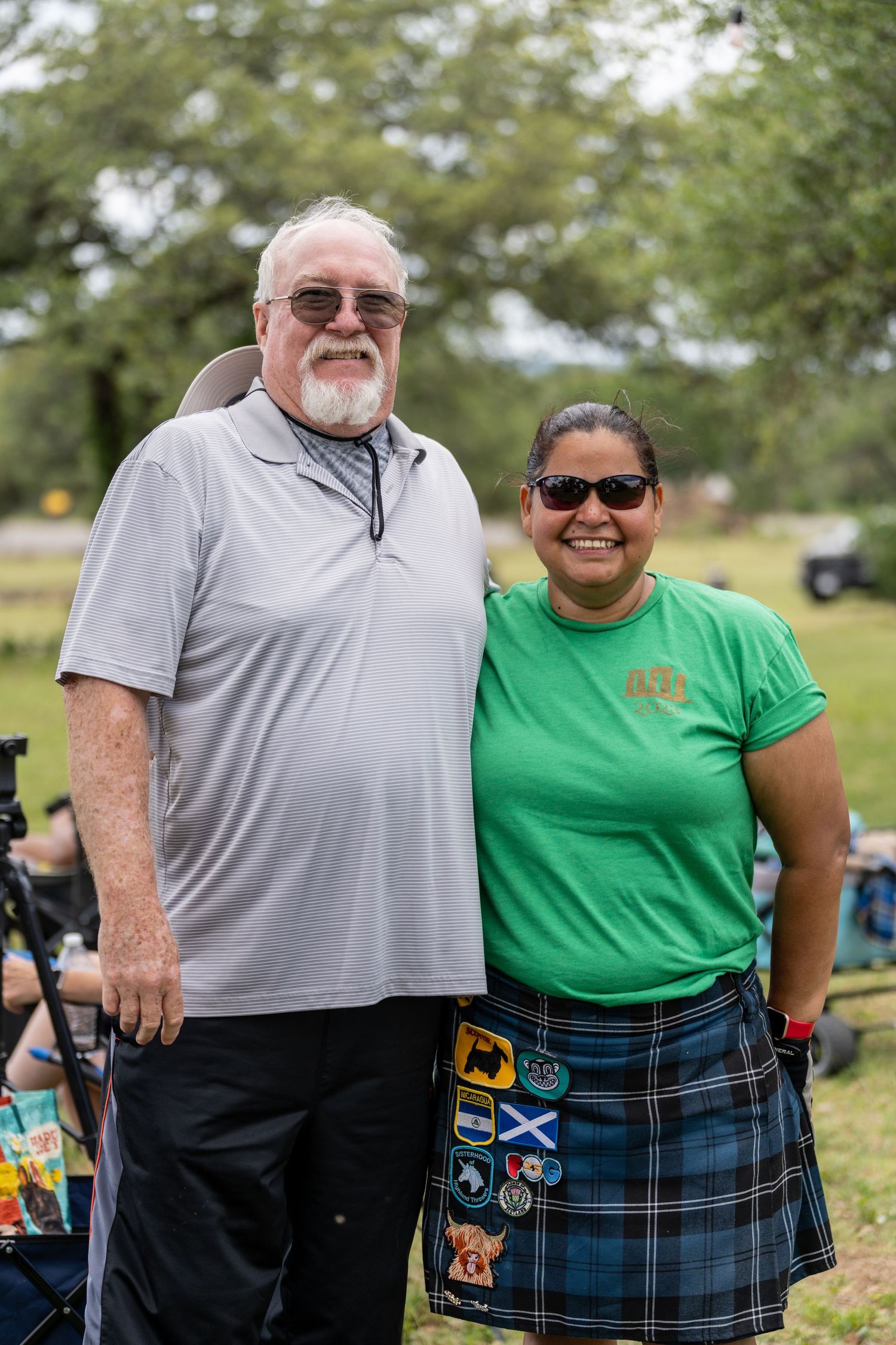 Two people smiling outdoors at a Scottish Highland Games event, woman wearing a tartan kilt with patches and green shirt.