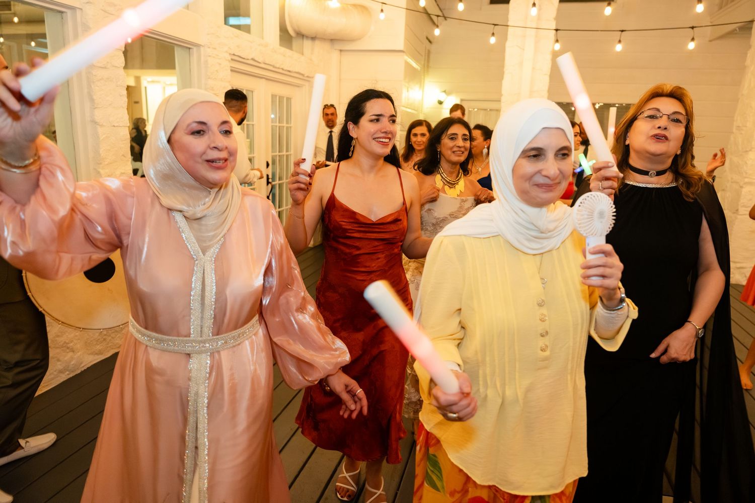 Women in elegant attire wave glow sticks at a festive indoor celebration event.