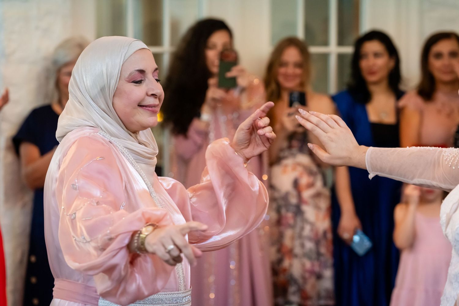 Woman in pink hijab dancing gracefully at a celebration, surrounded by guests in elegant attire capturing the moment.