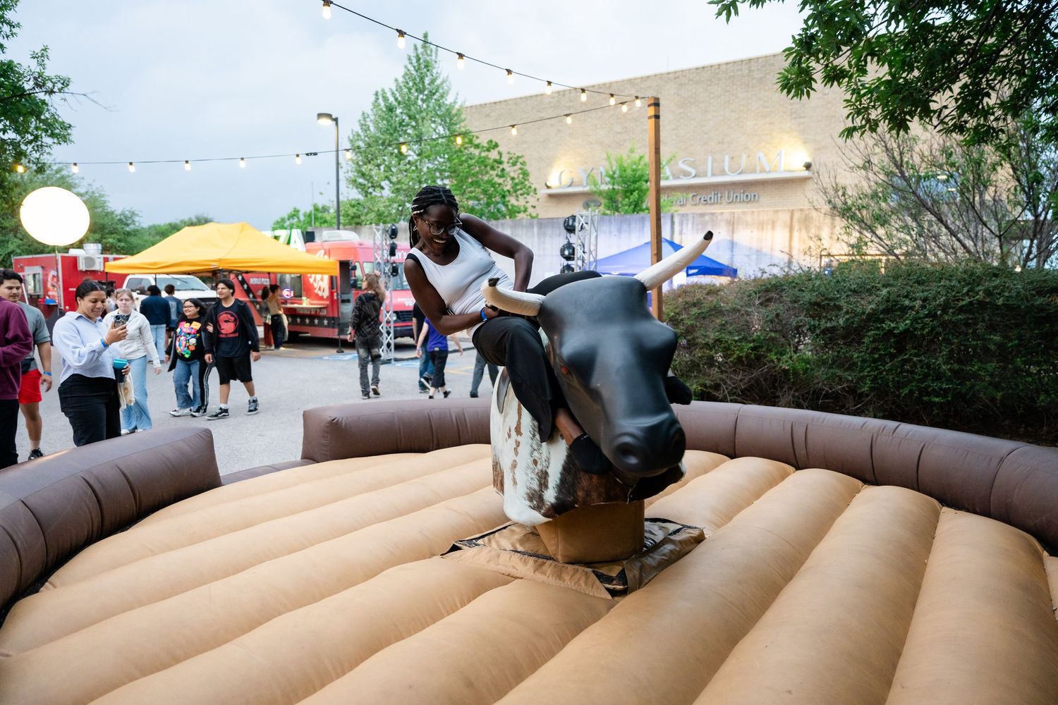 Person riding a mechanical bull at an outdoor festival event with colorful food vendors in the background.