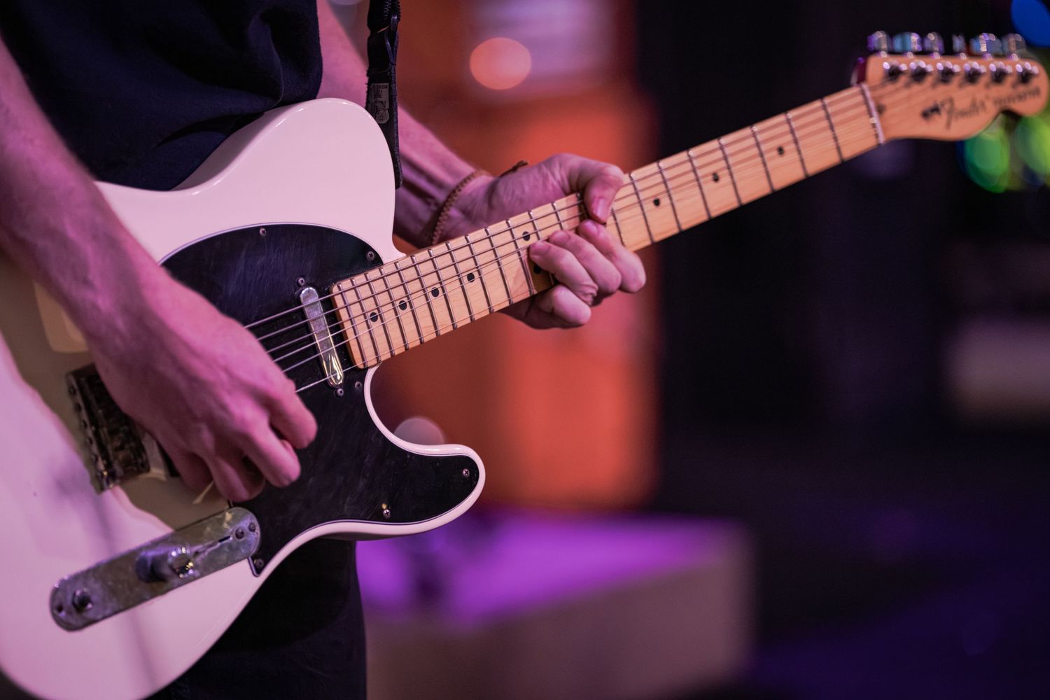 Musician playing a white Fender Telecaster electric guitar on a colorful lit stage during a live performance.