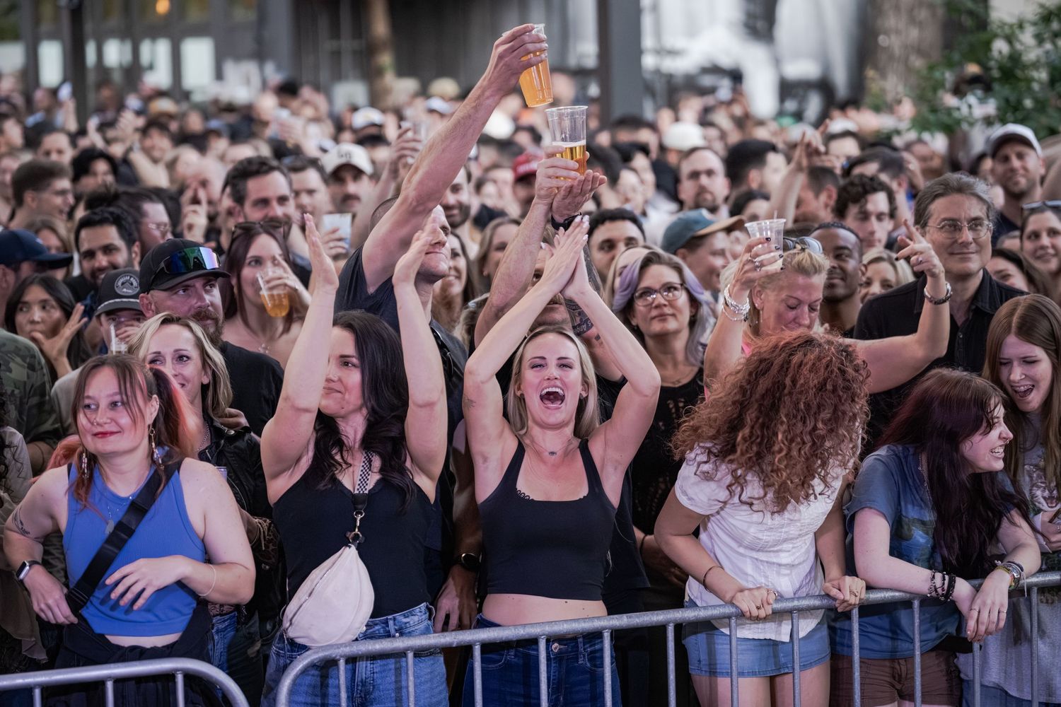 Excited crowd of fans cheering at an outdoor concert, with a woman raising her arms joyfully near a metal barrier.