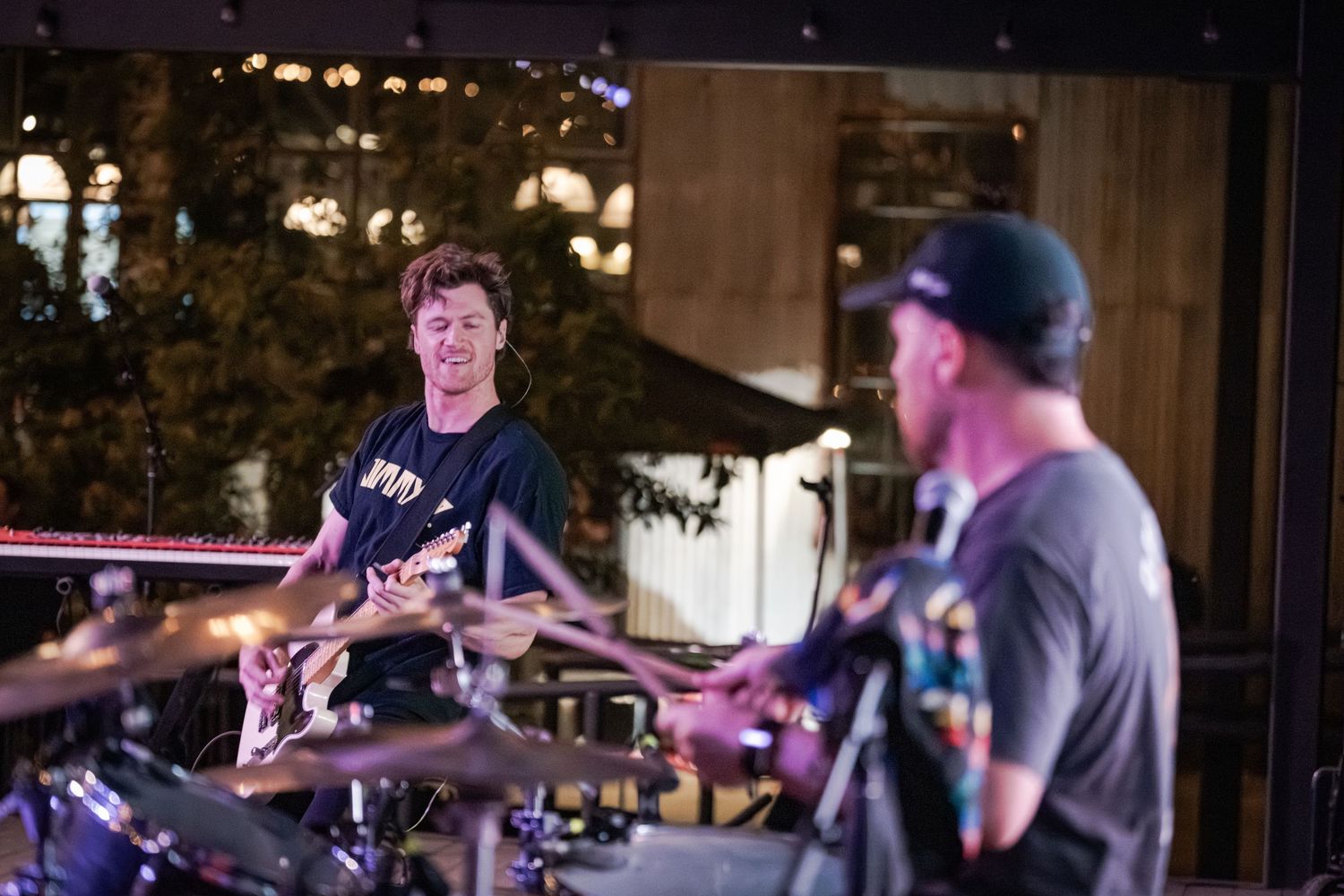 Two musicians perform live, one smiling guitarist and a drummer in a cap, on an outdoor stage at night.