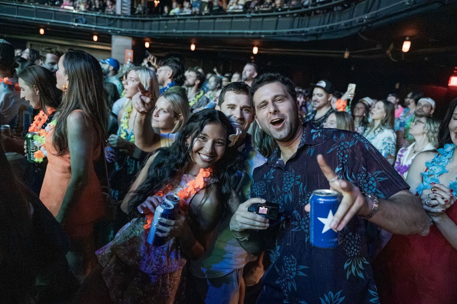 Three friends smile and hold drinks at a lively tropical-themed party with a crowded venue in the background.