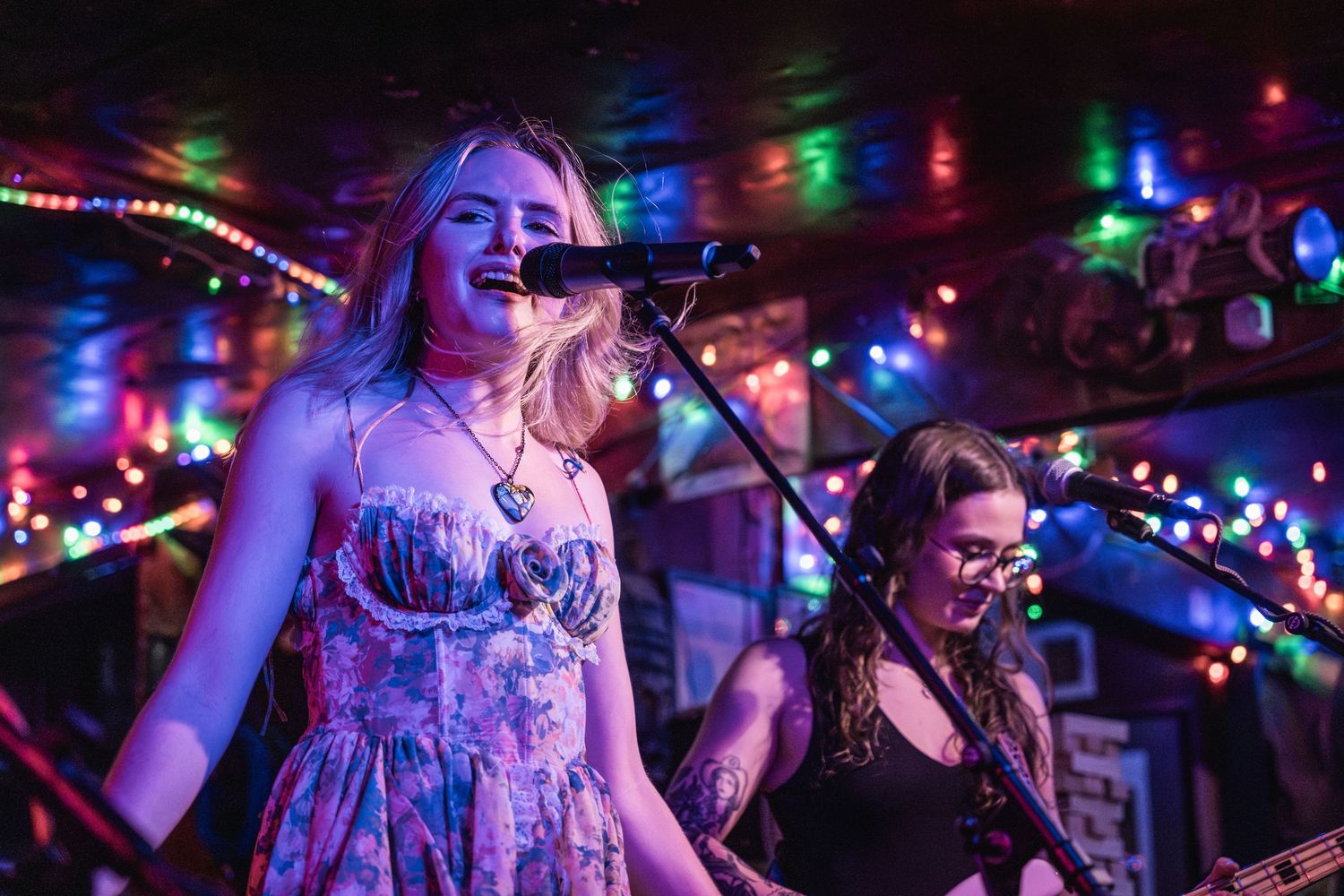 Musicians perform on stage at a small venue with colorful string lights creating a vibrant nightclub atmosphere.