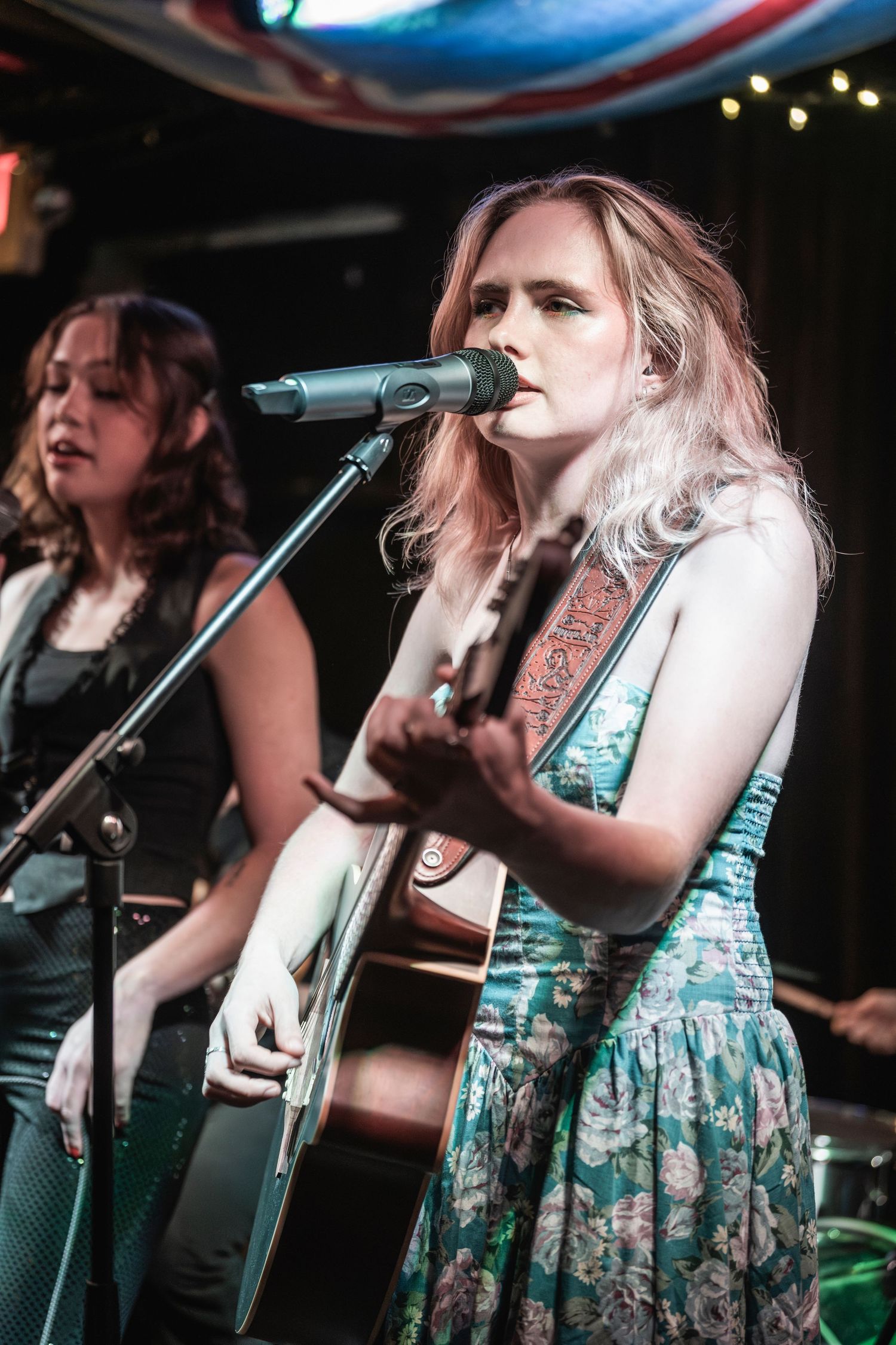 A musician in a floral dress performs on stage with a guitar under venue lighting.