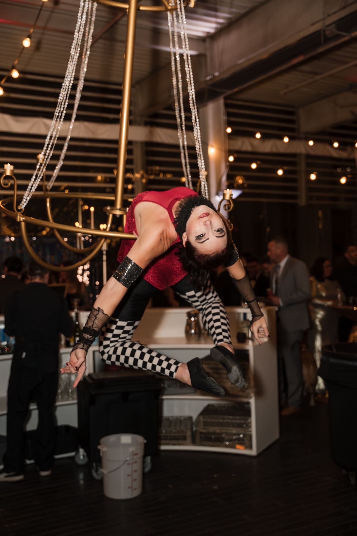 Four sequential photos show an aerial performer in a checkered outfit hanging and spinning from suspended rings at an indoor event.