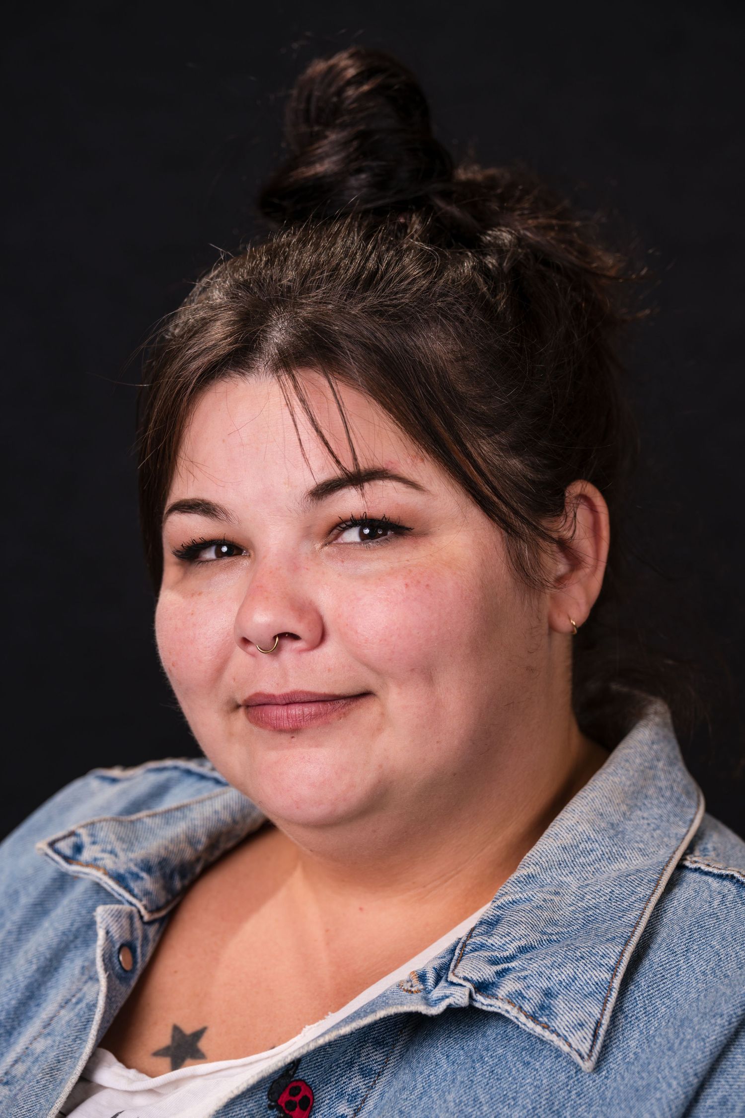 Portrait against dark background wearing denim jacket with hair in messy bun and natural makeup.