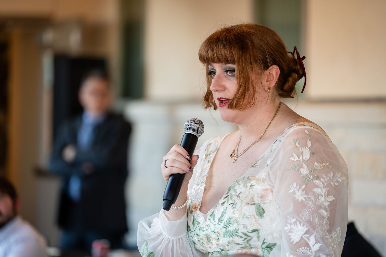 Someone in a white lace wedding dress delivers an emotional speech into a microphone during a reception.