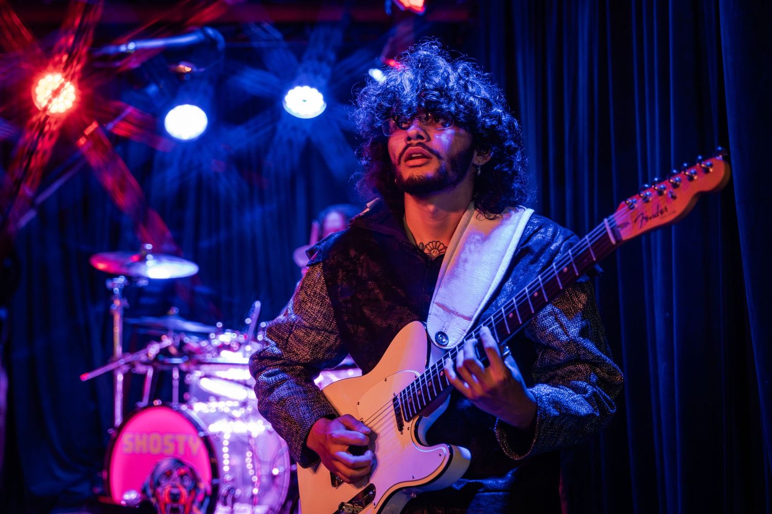 Musician plays electric guitar on dark stage illuminated by red and blue stage lights during live performance.