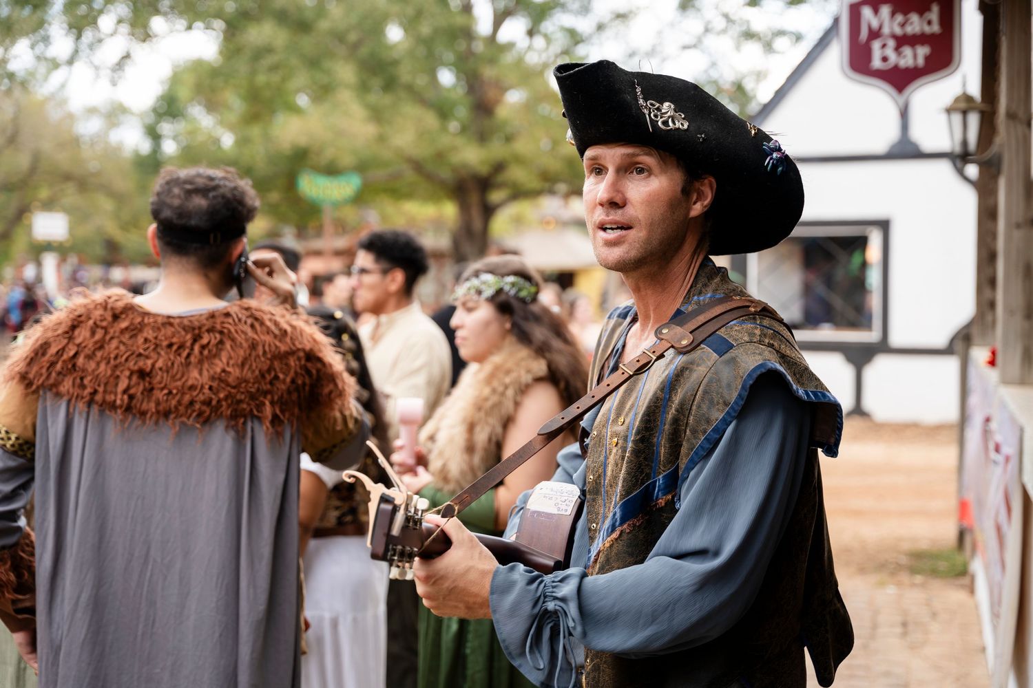 A person in medieval renaissance fair costume wearing a black hat and blue tunic stands among festival attendees.