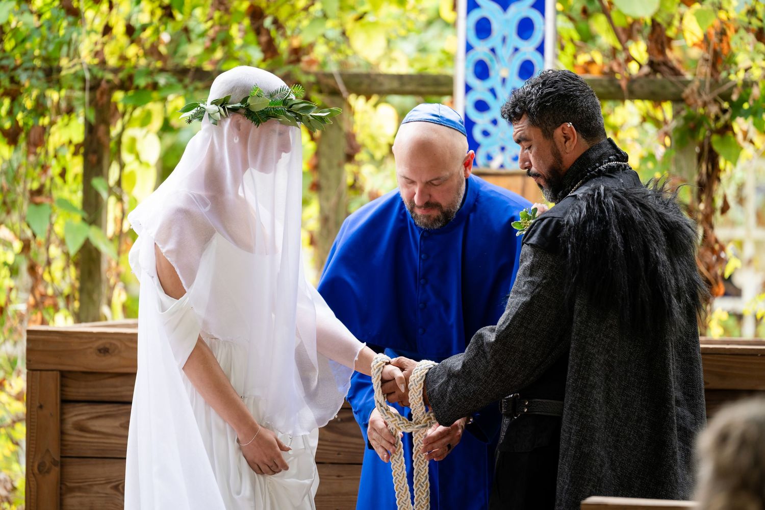 A couple exchanges vows during an outdoor garden wedding ceremony with a blue-robed officiant under a vine-covered pergola.