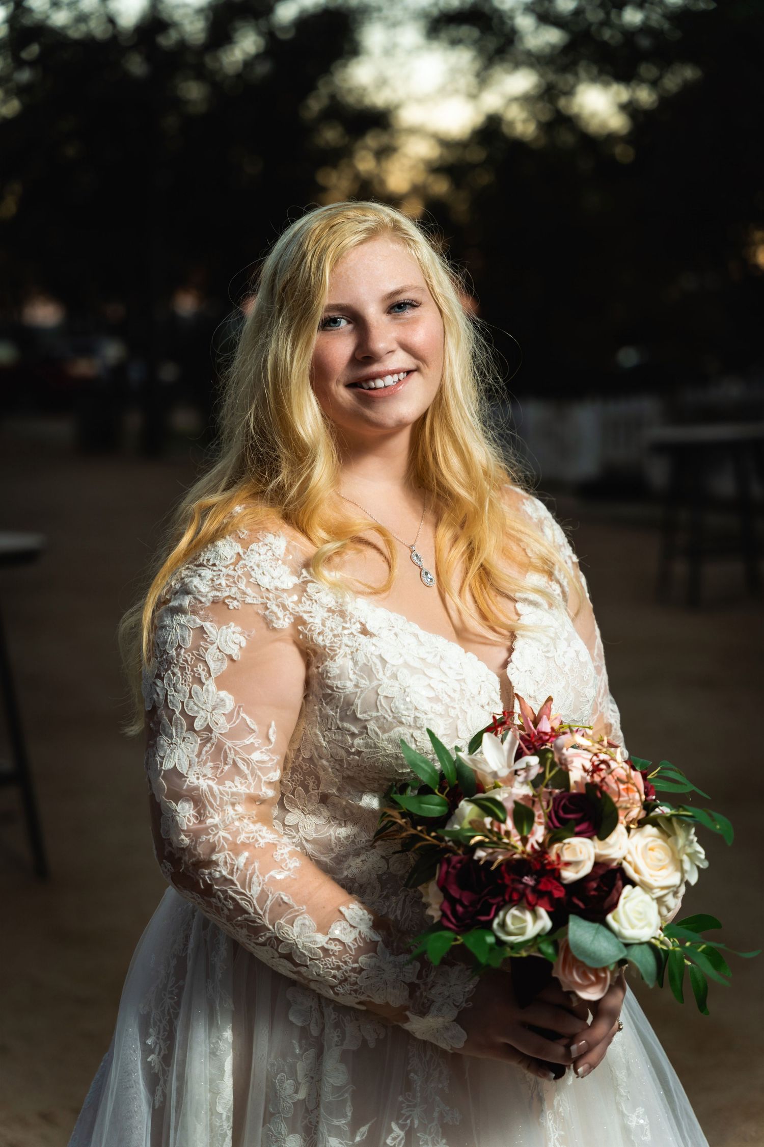 A bride in a white lace dress with long sleeves holds a burgundy and cream floral bouquet while smiling outdoors.