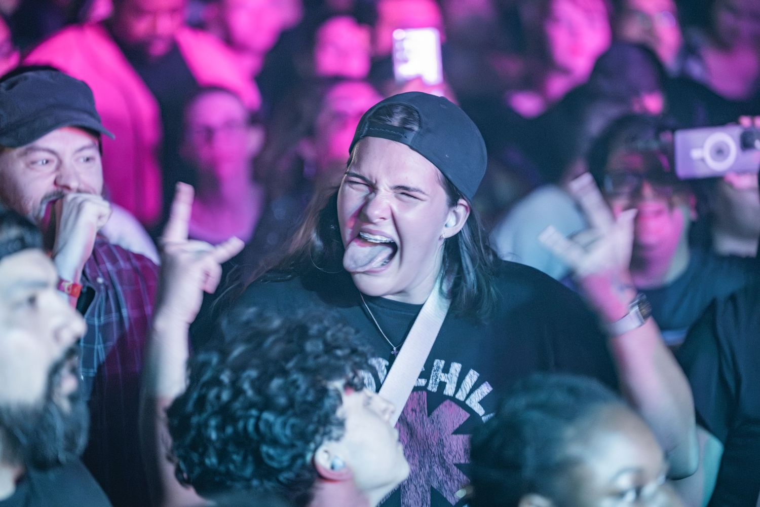 Concert audience members smile and cheer while bathed in vibrant purple lighting at a live music event.