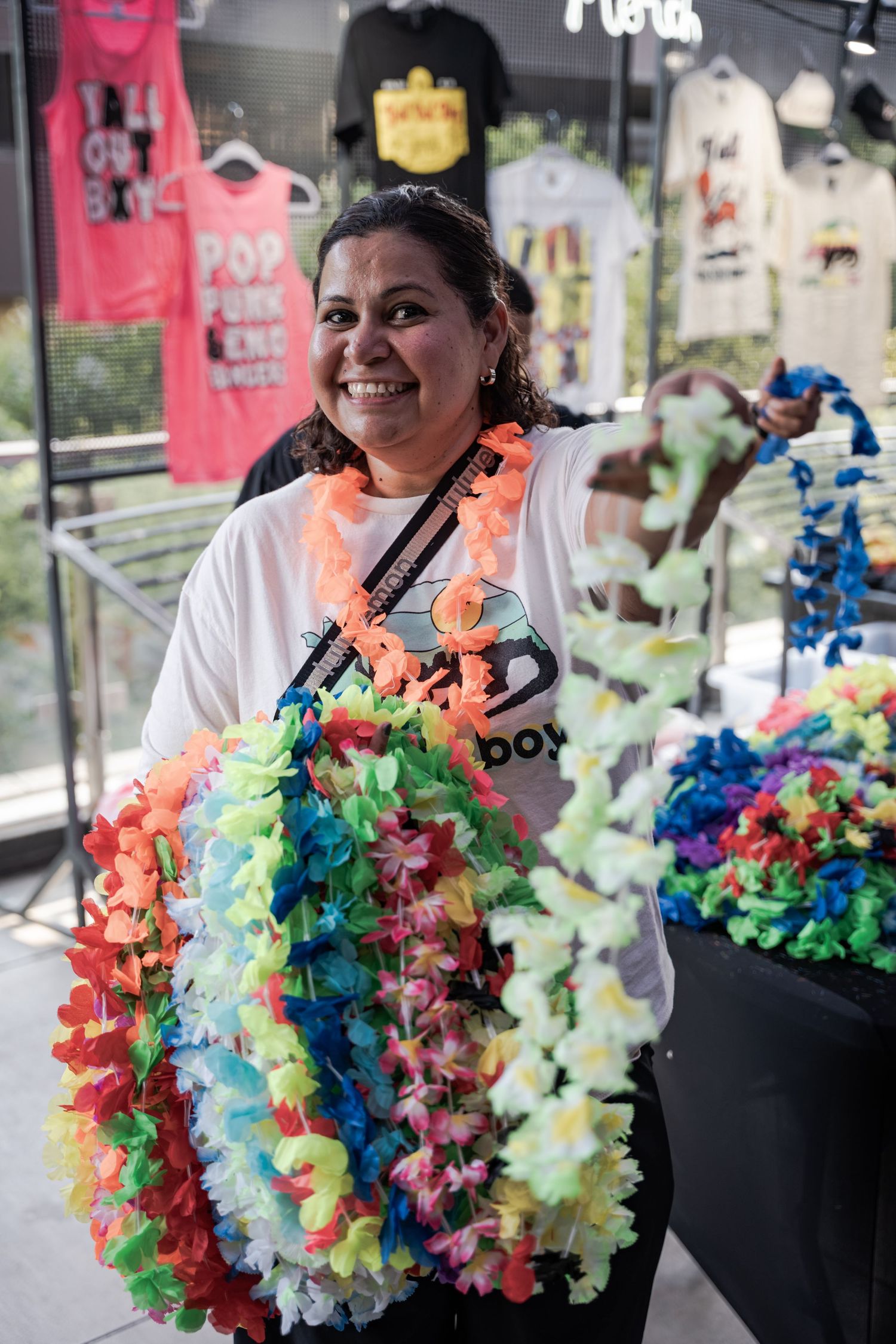 Smiling woman vendor holding colorful Hawaiian leis at an outdoor market with t-shirts displayed in background.