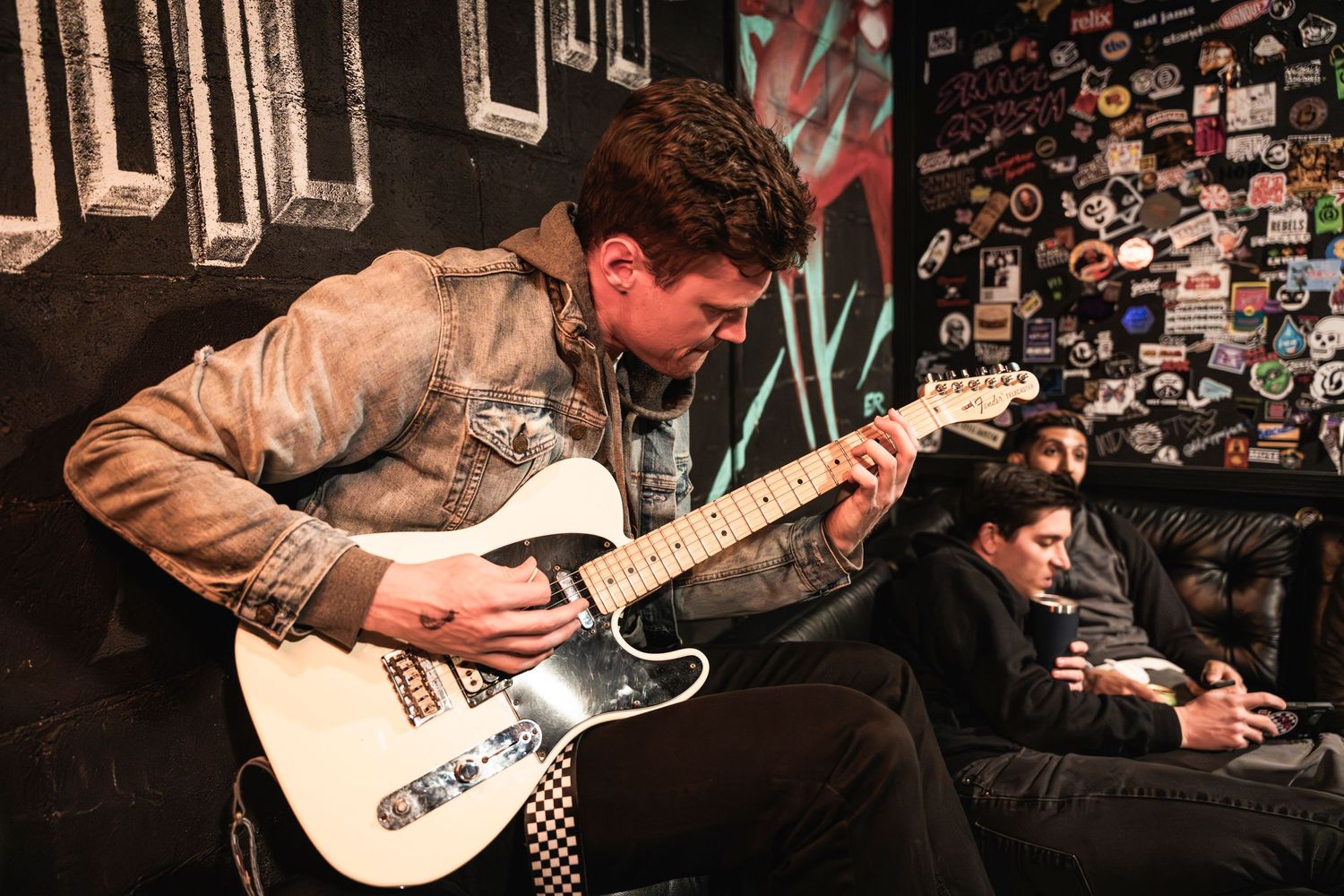 Musician playing white electric guitar in a dimly lit venue with decorative wall art.