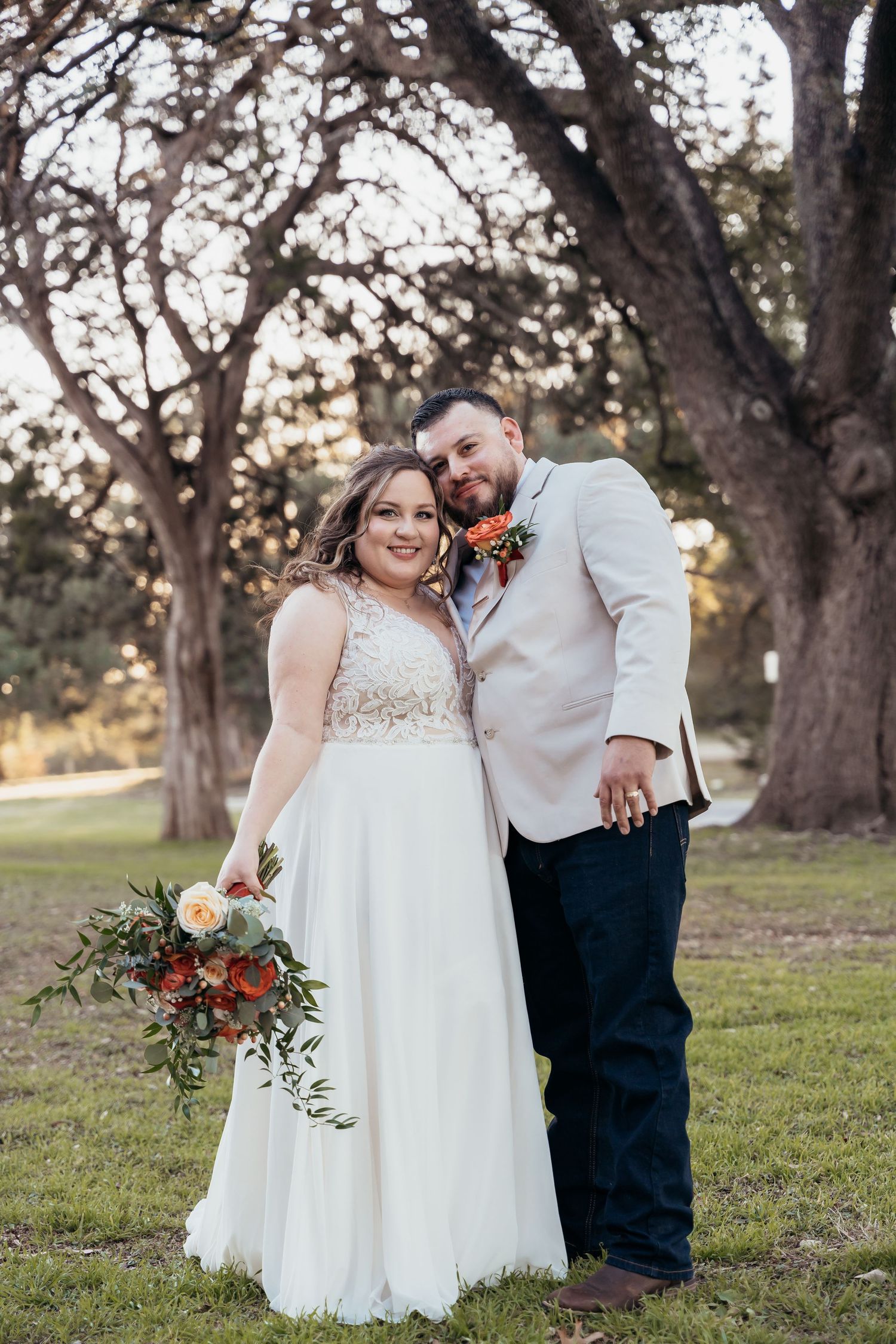 A couple poses on grass beneath towering oak trees during golden hour.