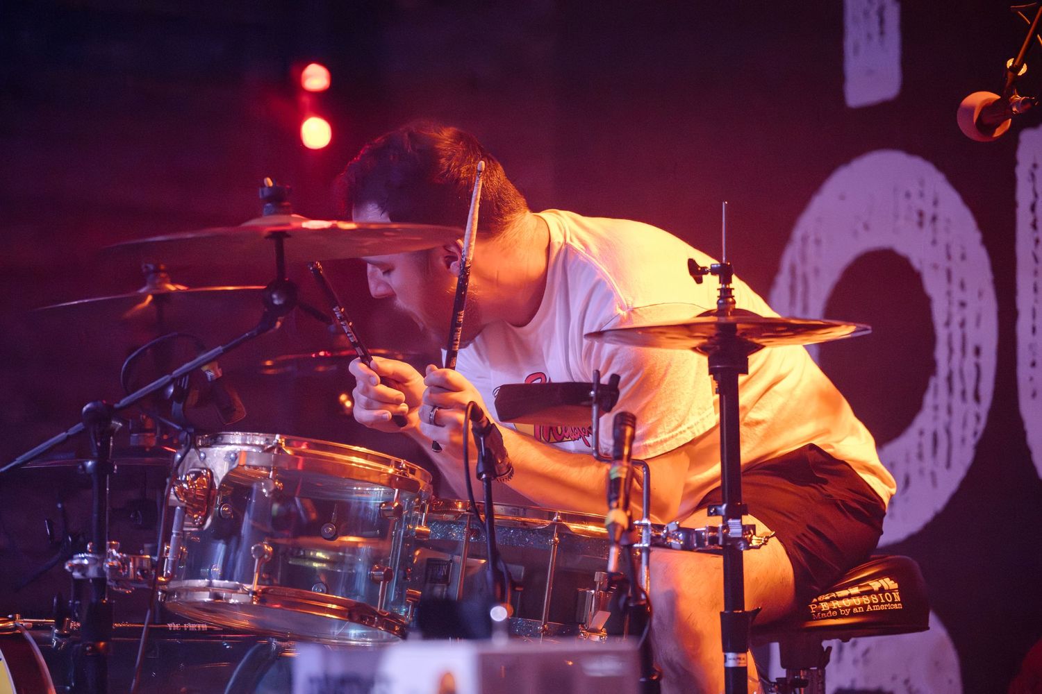 A drummer plays intensely on a drum kit under red stage lighting during a live performance.