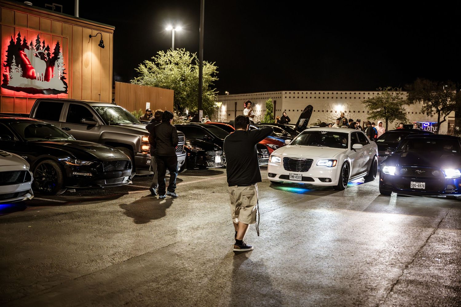 Car enthusiasts gathered in parking lot at night for automotive meet.
