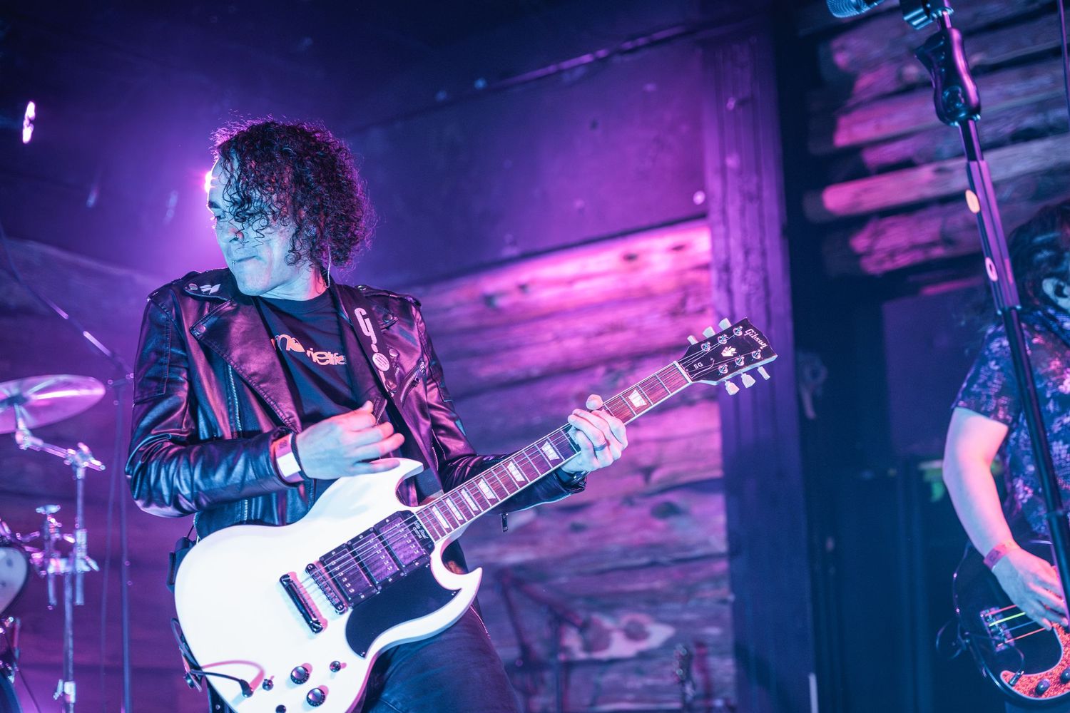 Guitarist playing white guitar on stage with purple backlit wood panel wall.