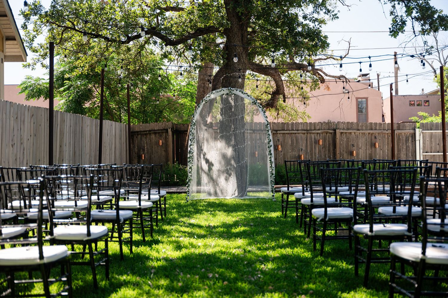 Outdoor wedding ceremony setup with white chairs and archway under a large tree.