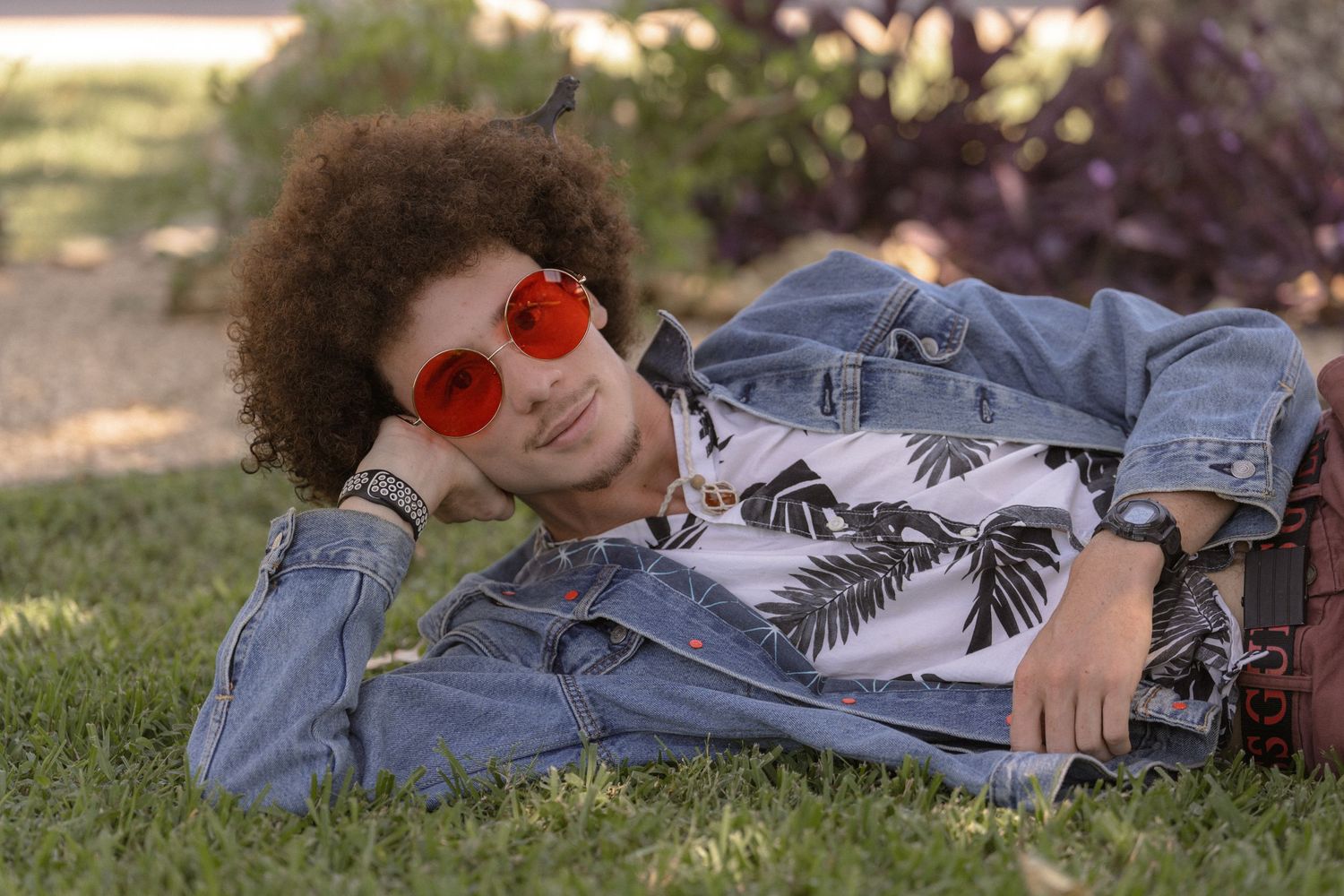 Person with afro hair relaxing on grass wearing red sunglasses and tropical print shirt.