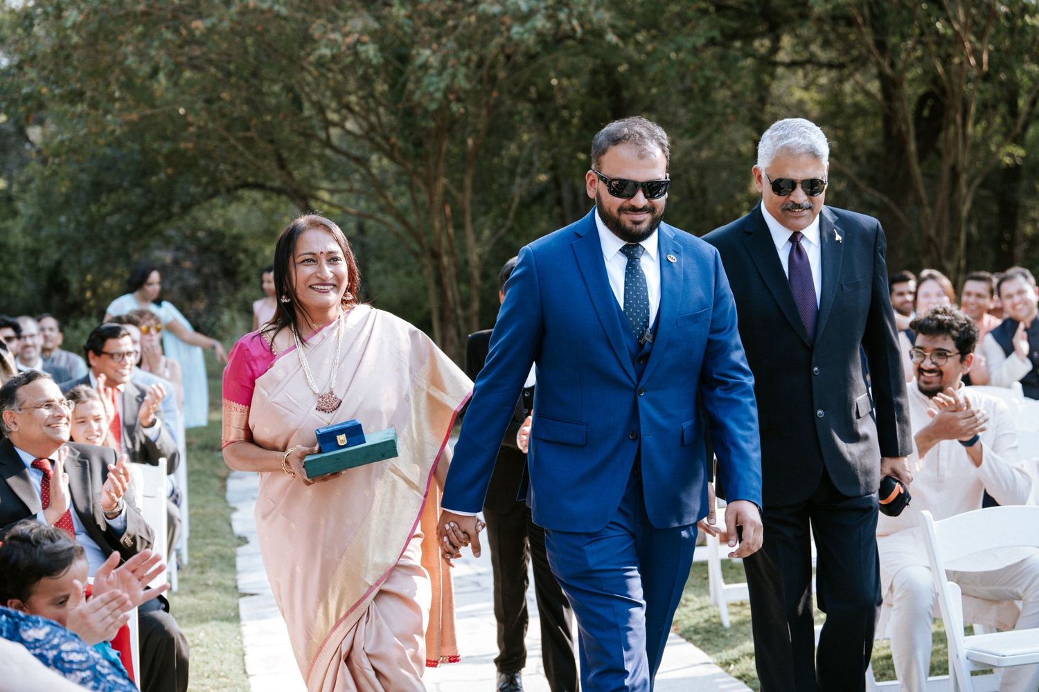 Wedding party members walking down an outdoor aisle surrounded by guests in white chairs.