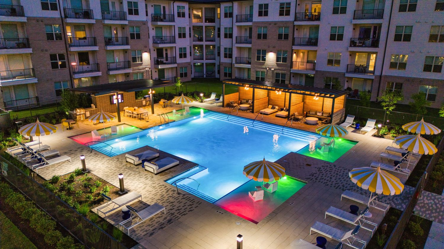 Modern apartment complex swimming pool with colorful LED lighting and lounge areas at night.