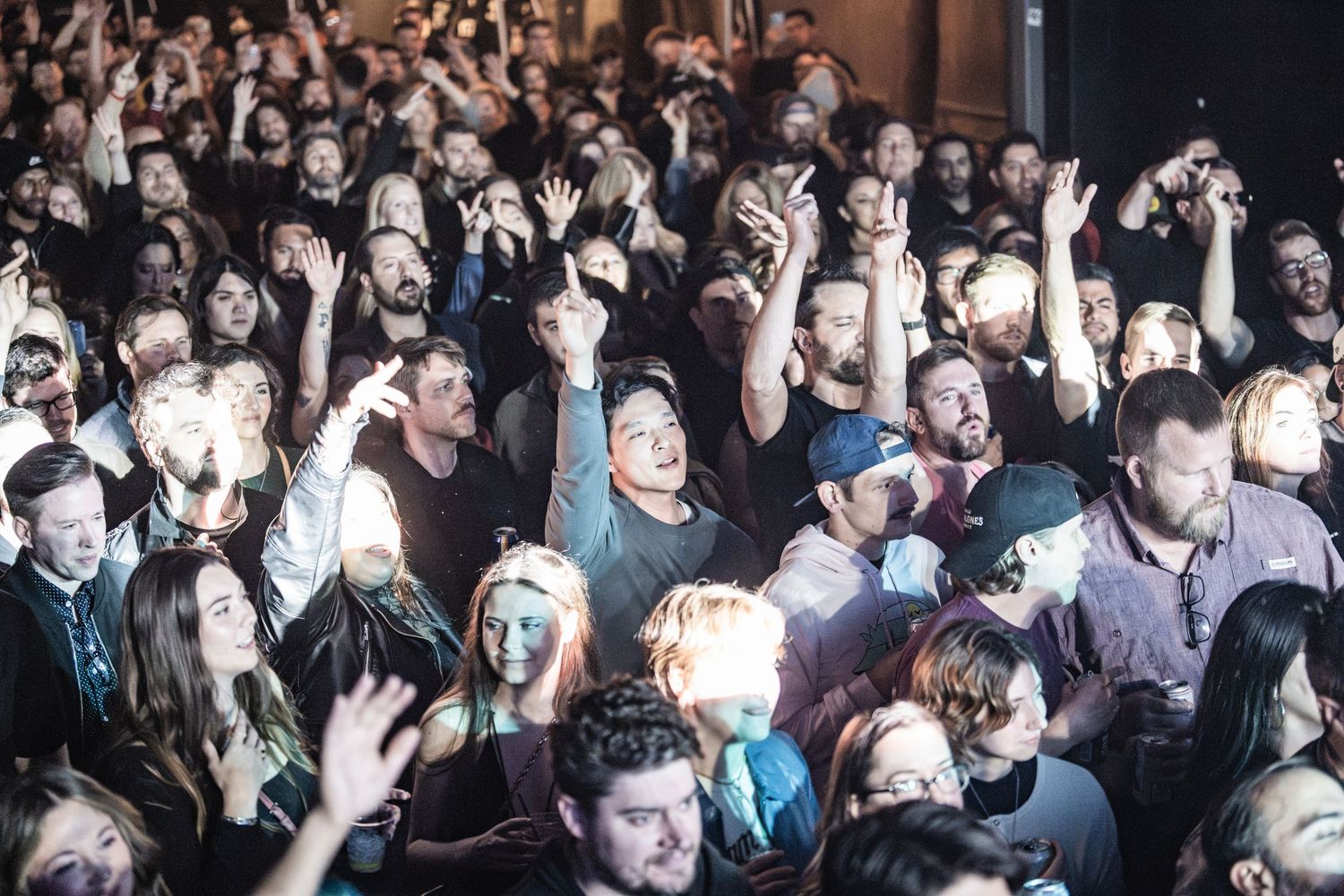 Large crowd of concert attendees packed together at live music event with illuminated faces in dark venue.