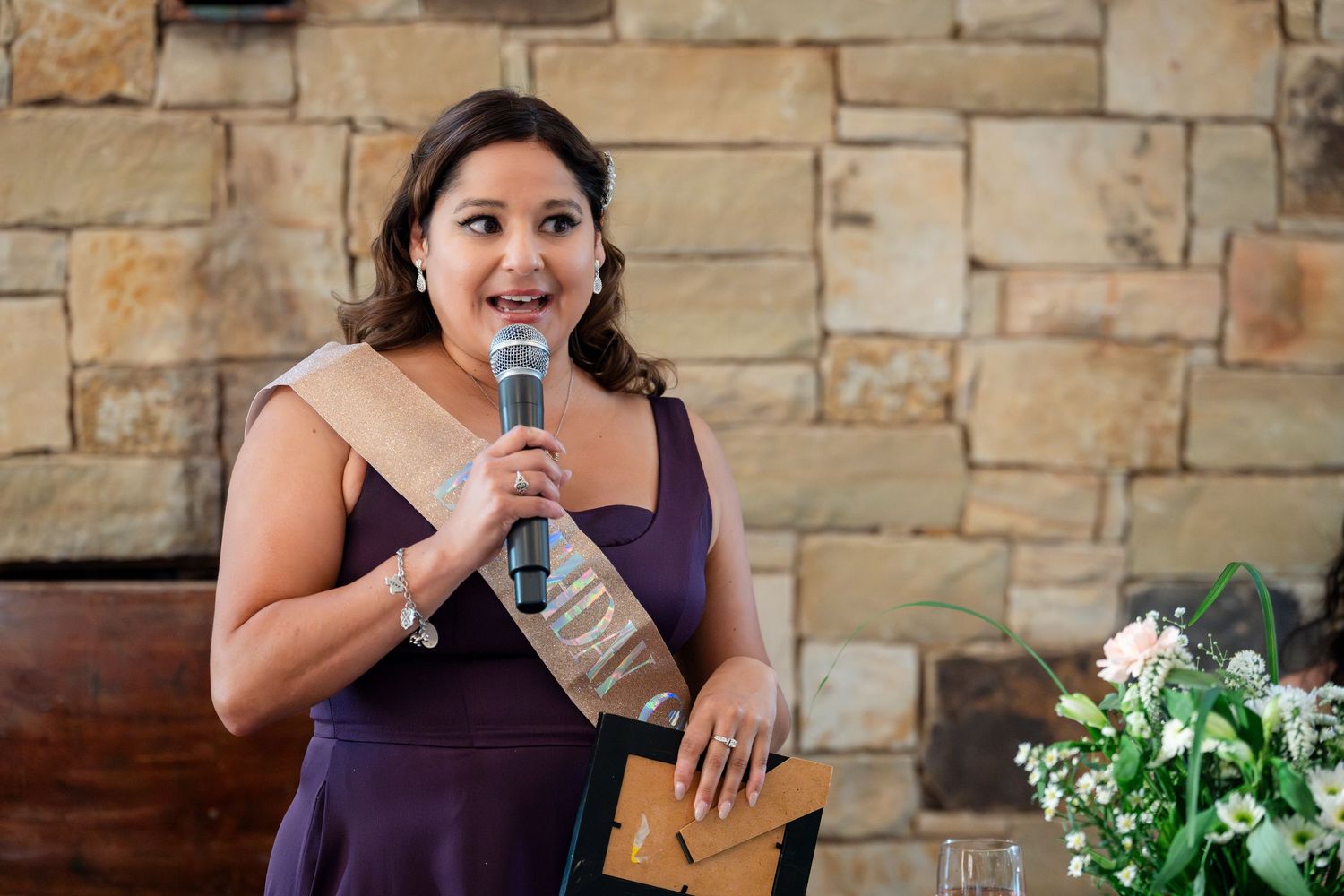 A speaker in a purple dress holds a microphone while speaking at an event with a stone wall backdrop and floral decor.