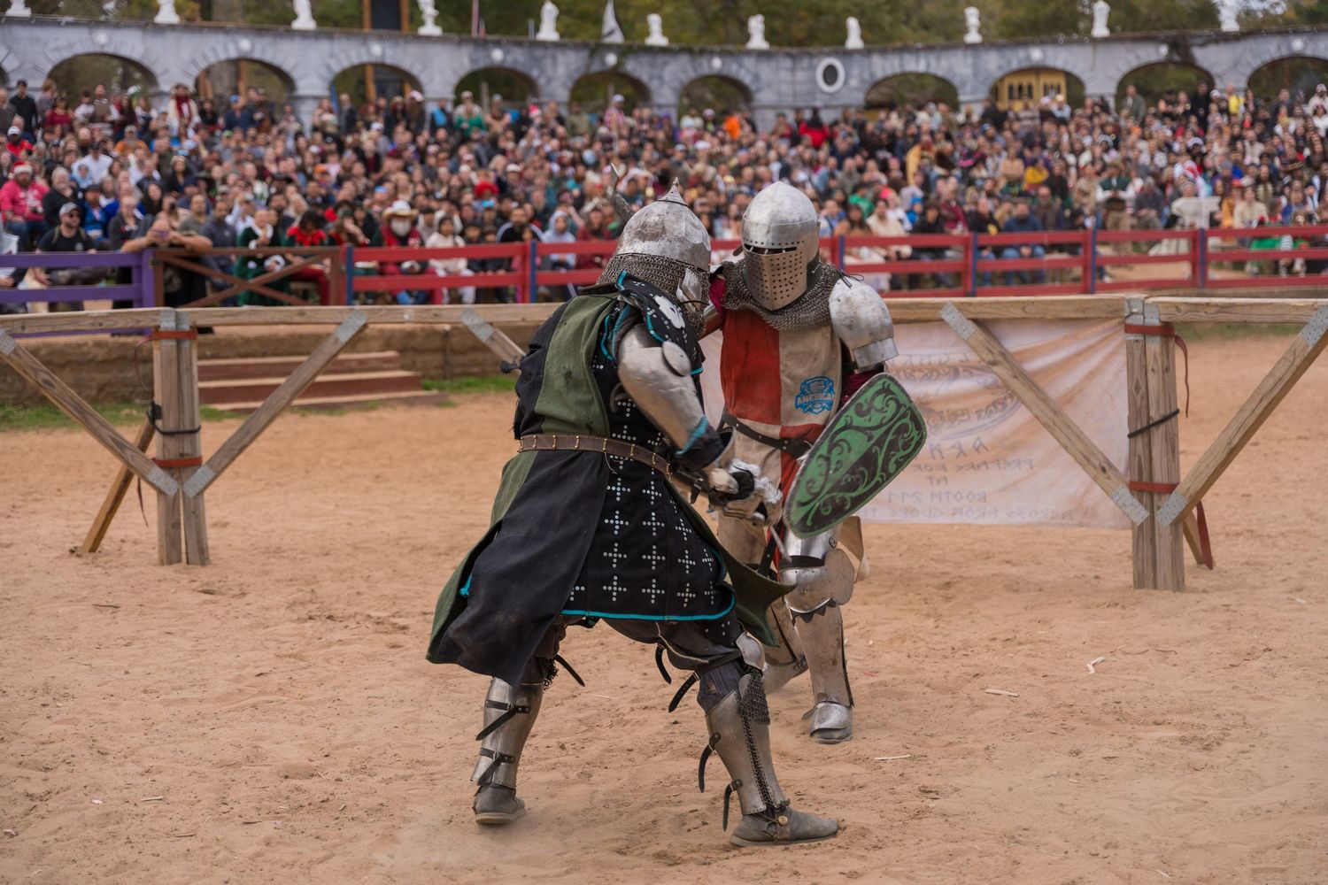 Armored medieval knights duel with swords in a sand arena before a large crowd.