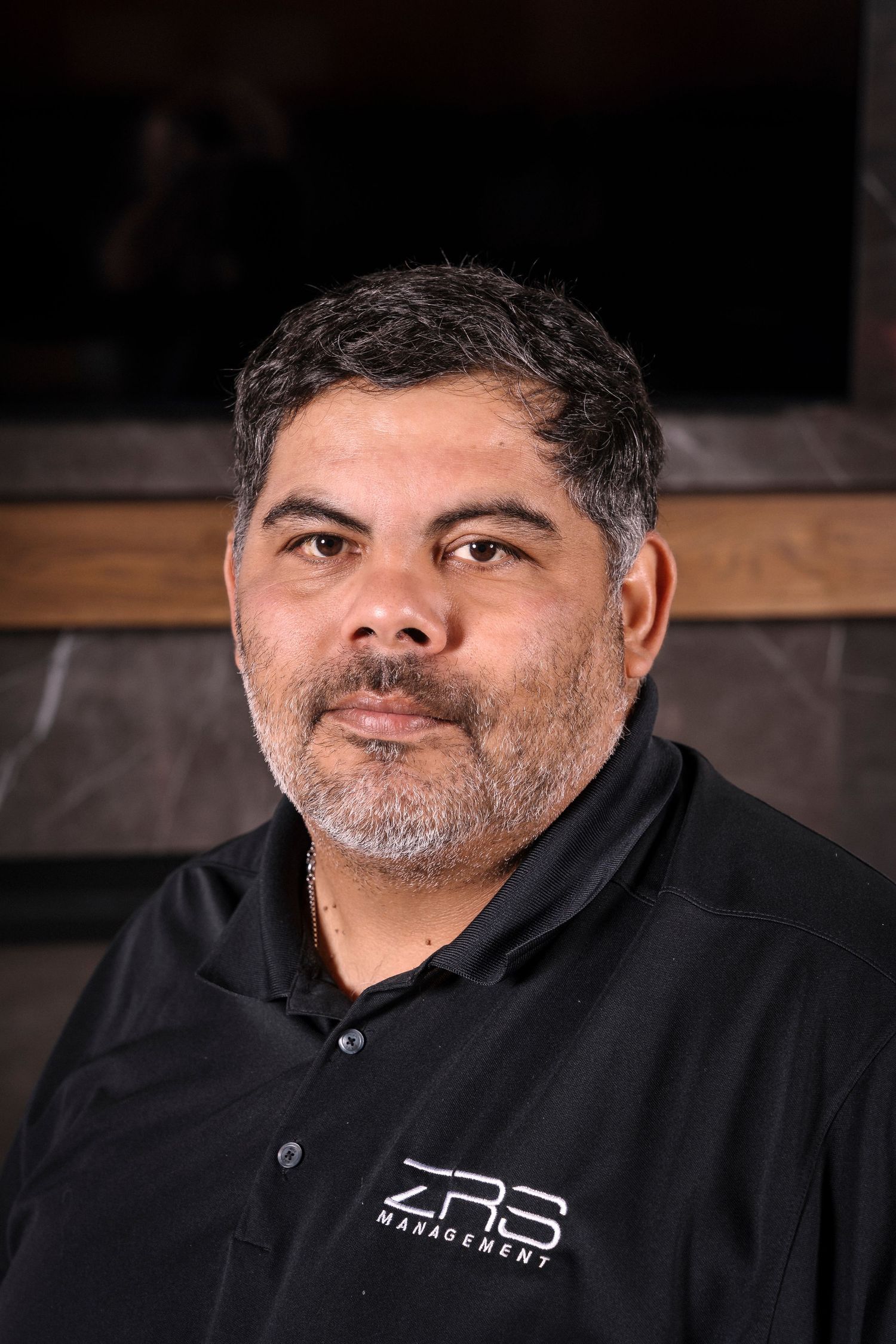 Professional studio portrait of someone in a black polo shirt against a dark background with wooden accents.