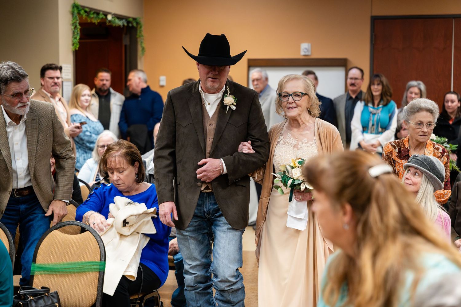 Wedding guests gather in a reception hall with someone wearing a cowboy hat alongside a person in a champagne-colored dress.