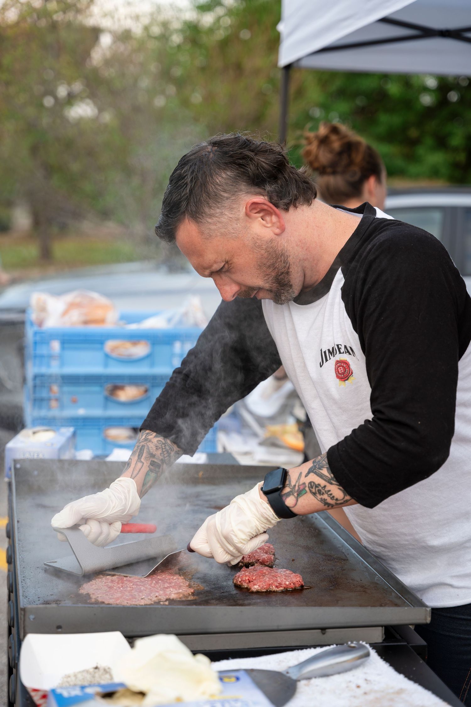 Chef on outdoor grill preparing hamburger patties at catering event with gloved hands and meat prep station.