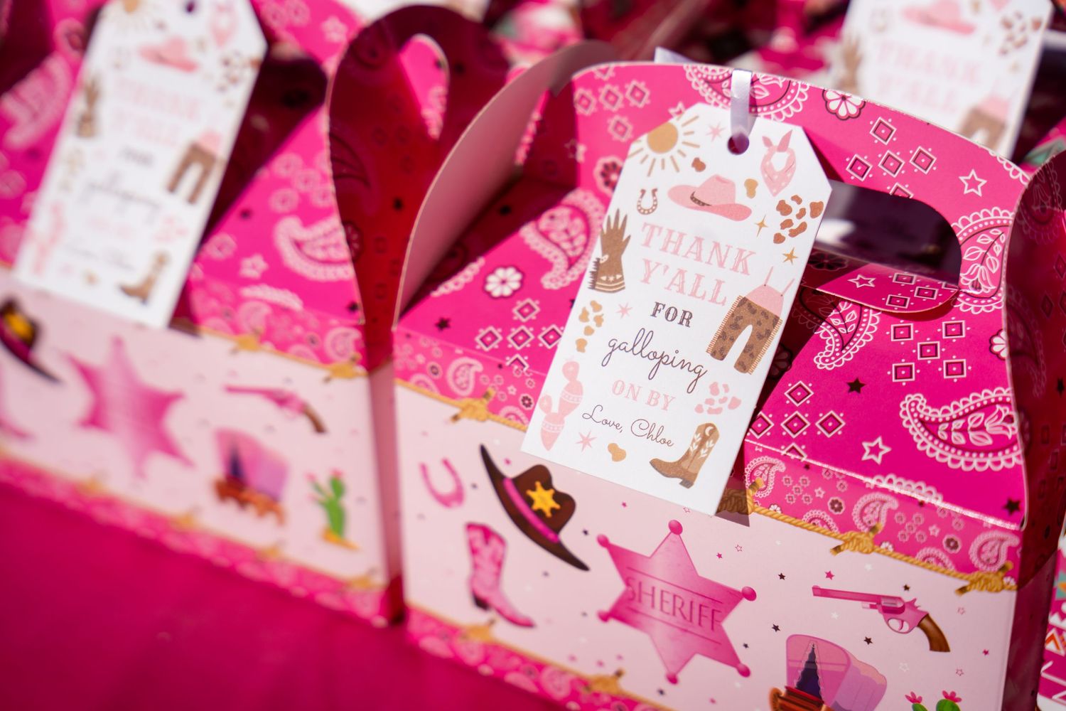 Pink party favor boxes with gift tags and swirling designs displayed in a row at an event.