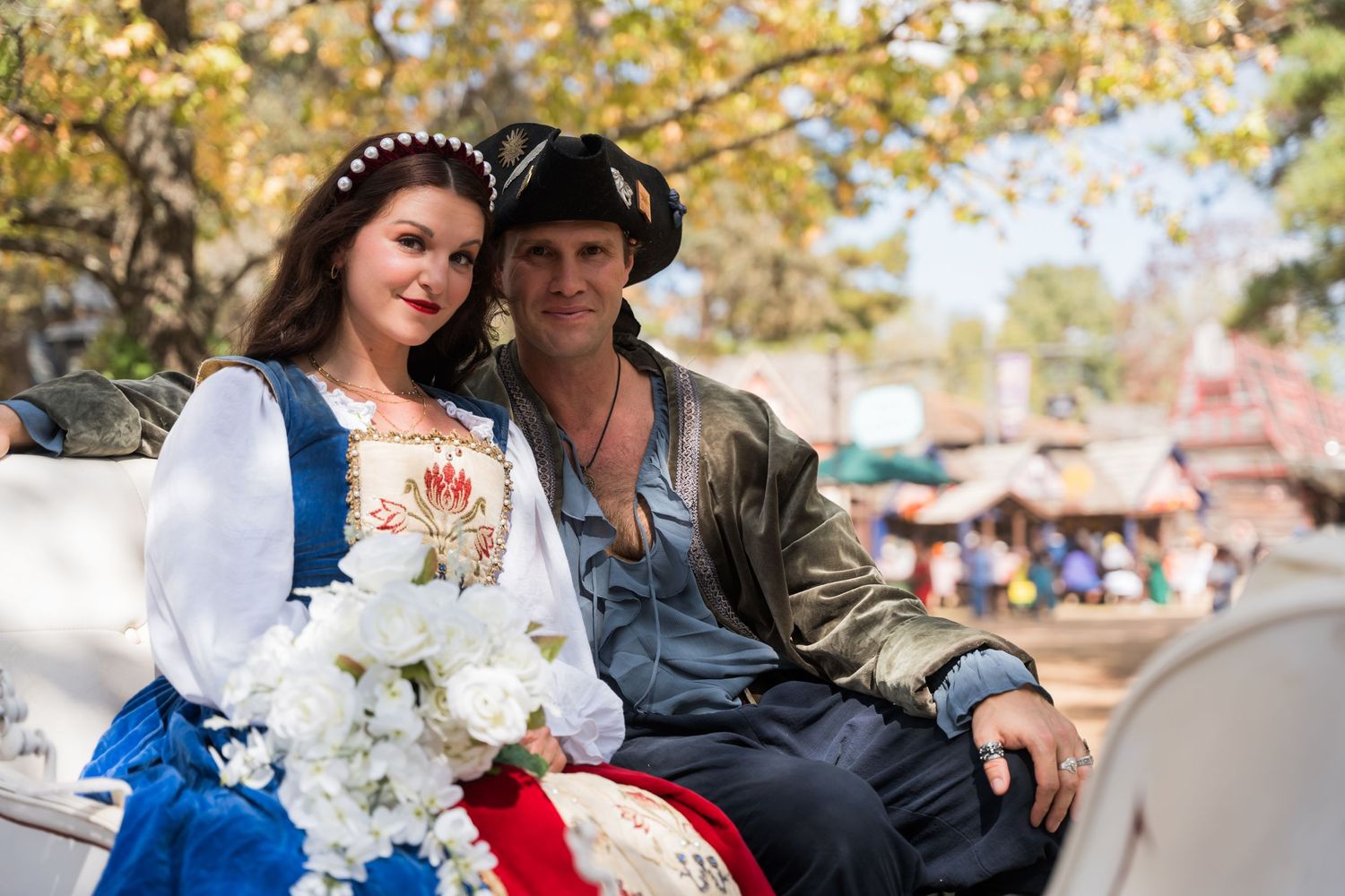 Couple in renaissance fair attire pose together on wooden bench with flowers and autumn trees in background.