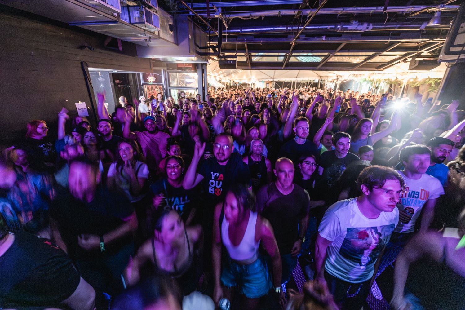 Large concert crowd fills venue floor under purple lighting during live performance.