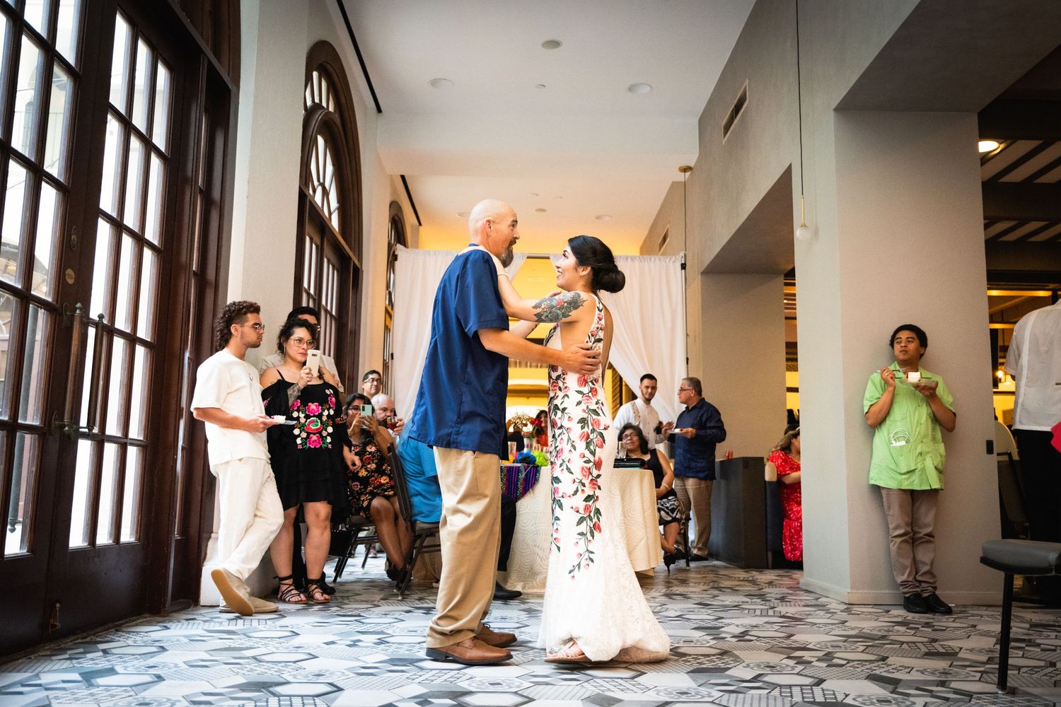Couples dance in an elegant hallway with patterned floors and large windows.