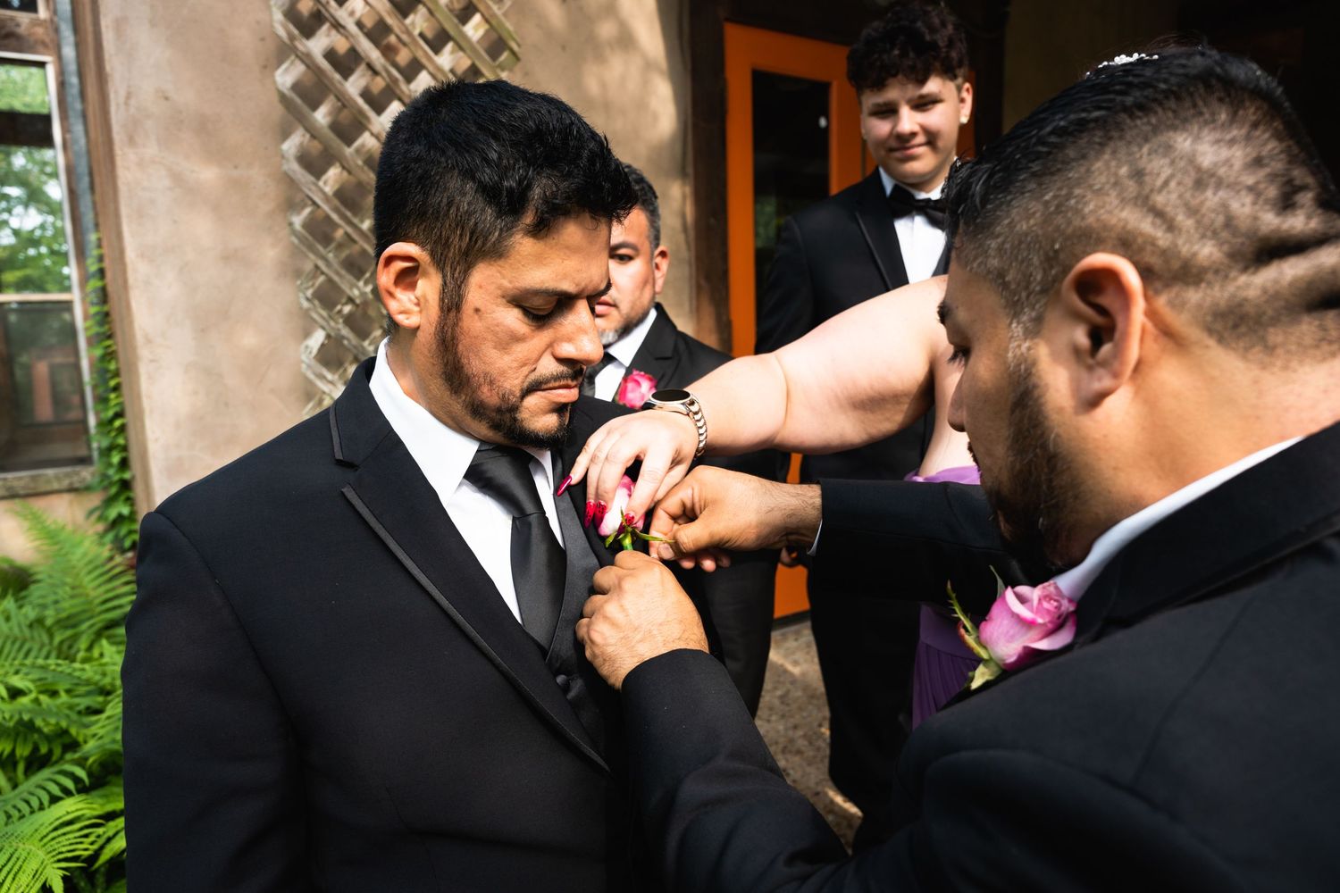 Groomsmen help adjust a boutonniere on a suit jacket at an outdoor wedding.