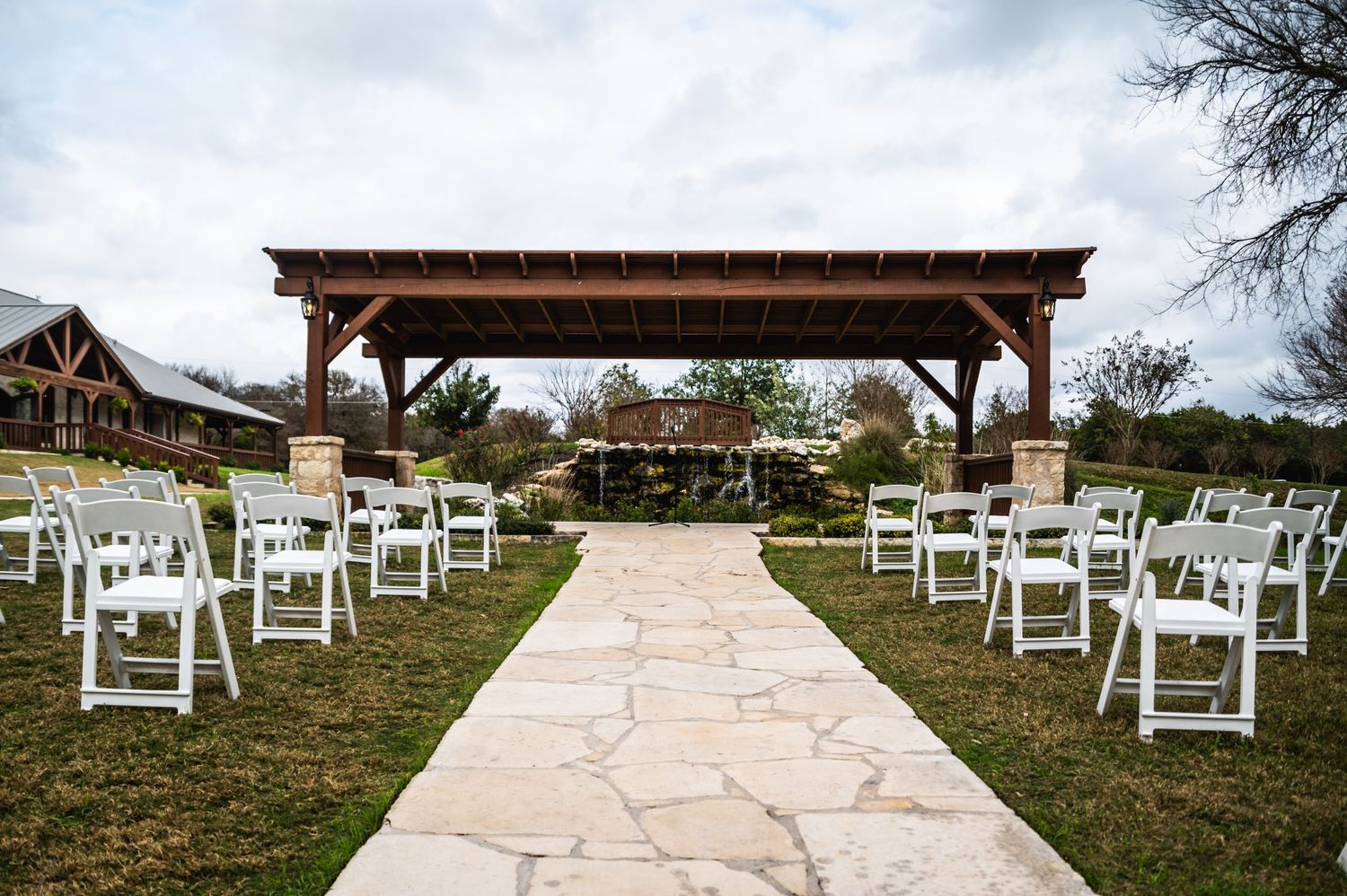 White chairs line a stone pathway leading to a wooden pavilion for an outdoor ceremony.
