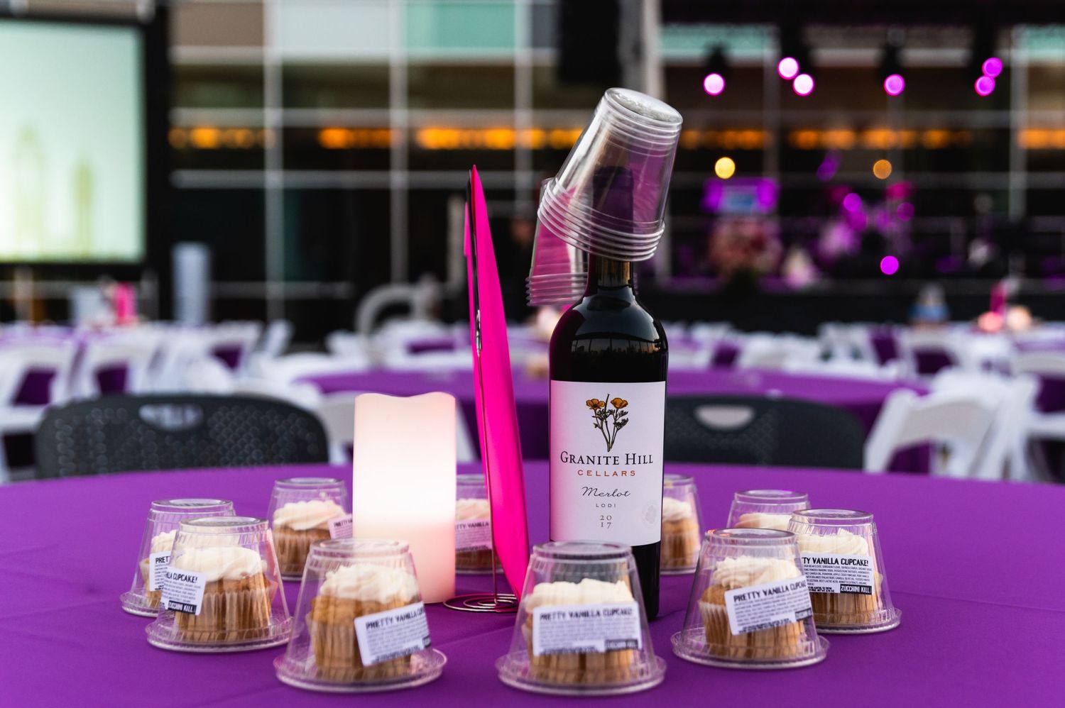 Wine bottle and dessert settings on purple tablecloth with decorative lighting.