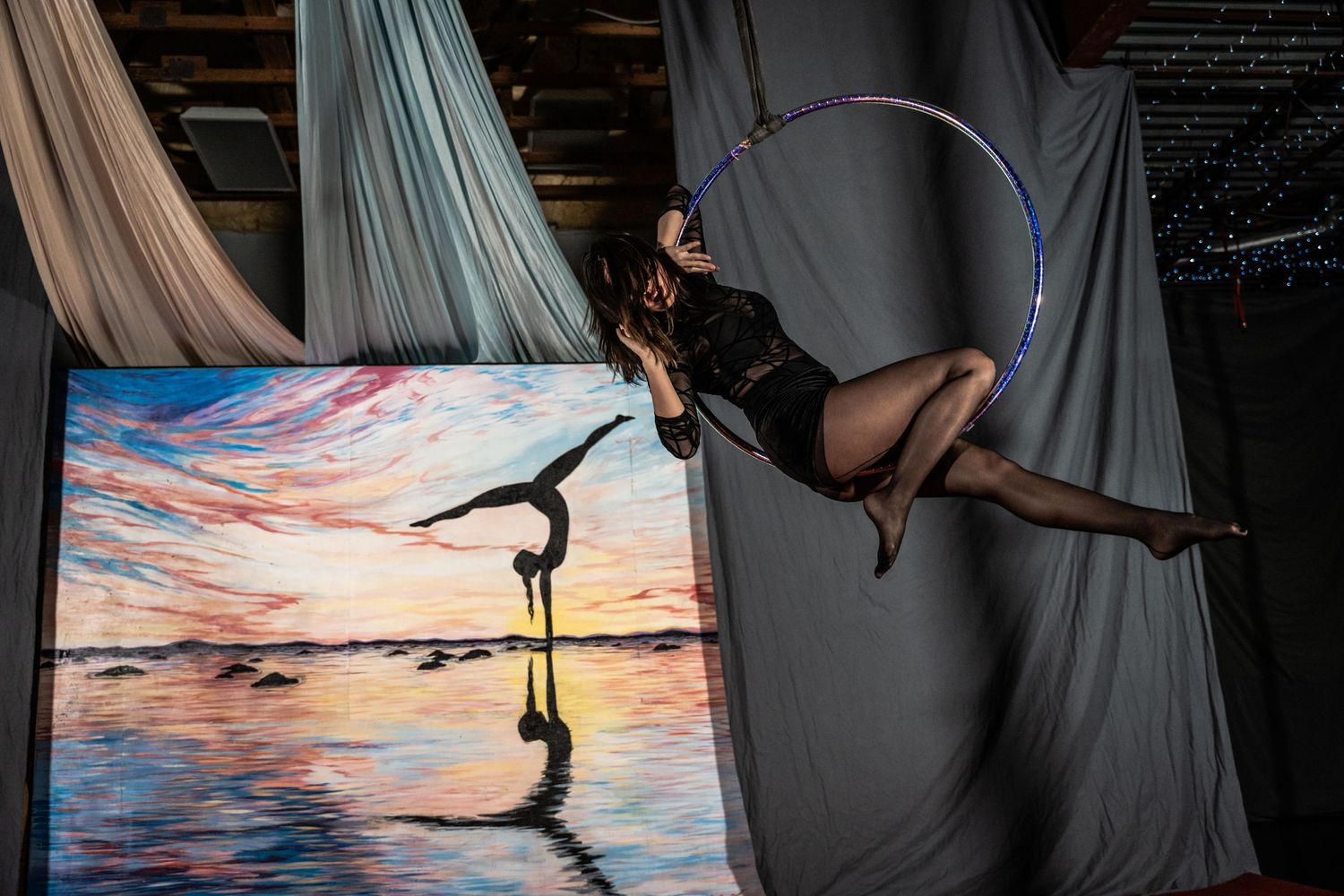 An aerial performer in black attire poses gracefully on a hoop against a sunset backdrop.