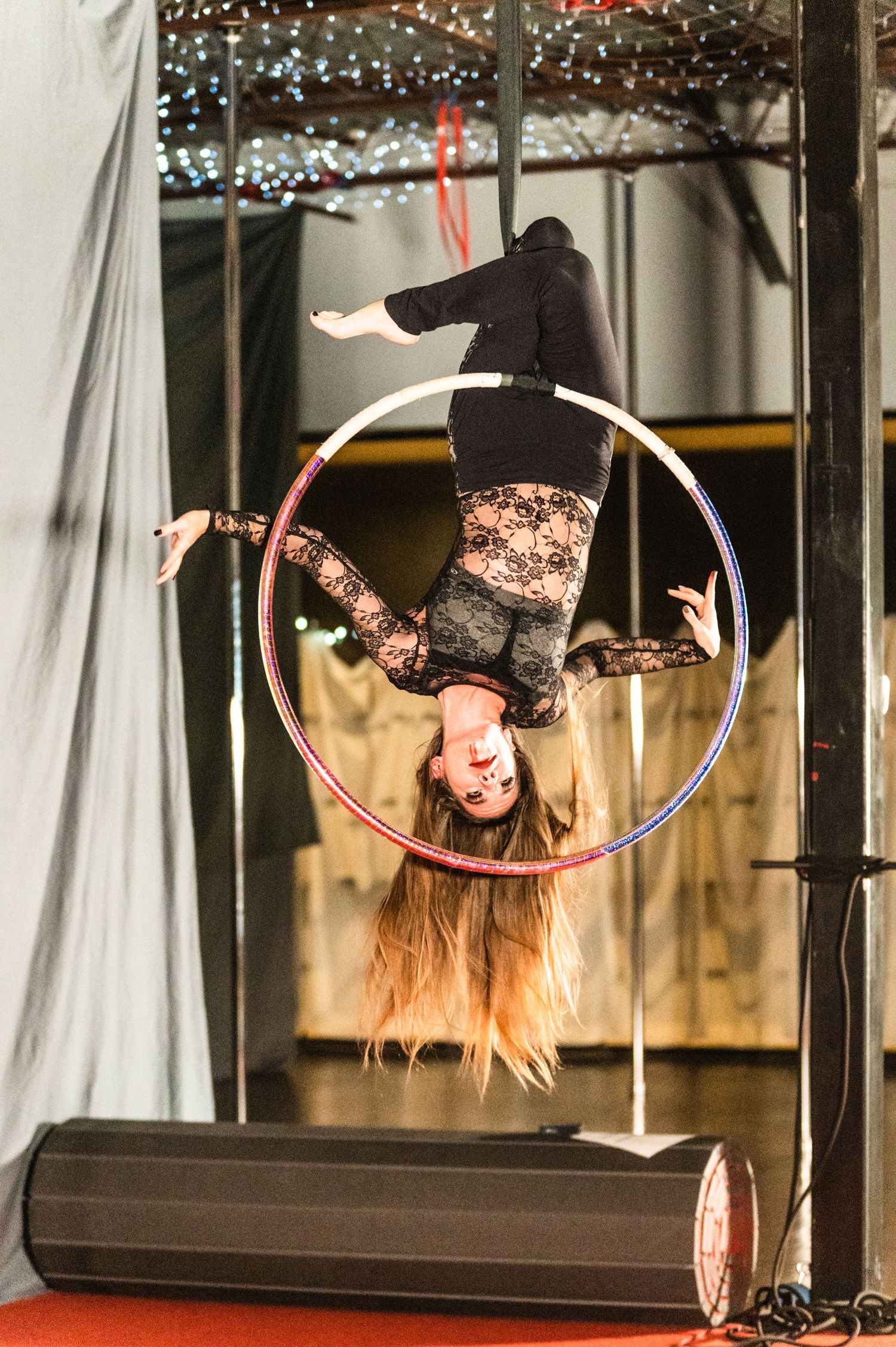 Performer hangs upside down in aerial hoop with long hair flowing beneath twinkle lights.